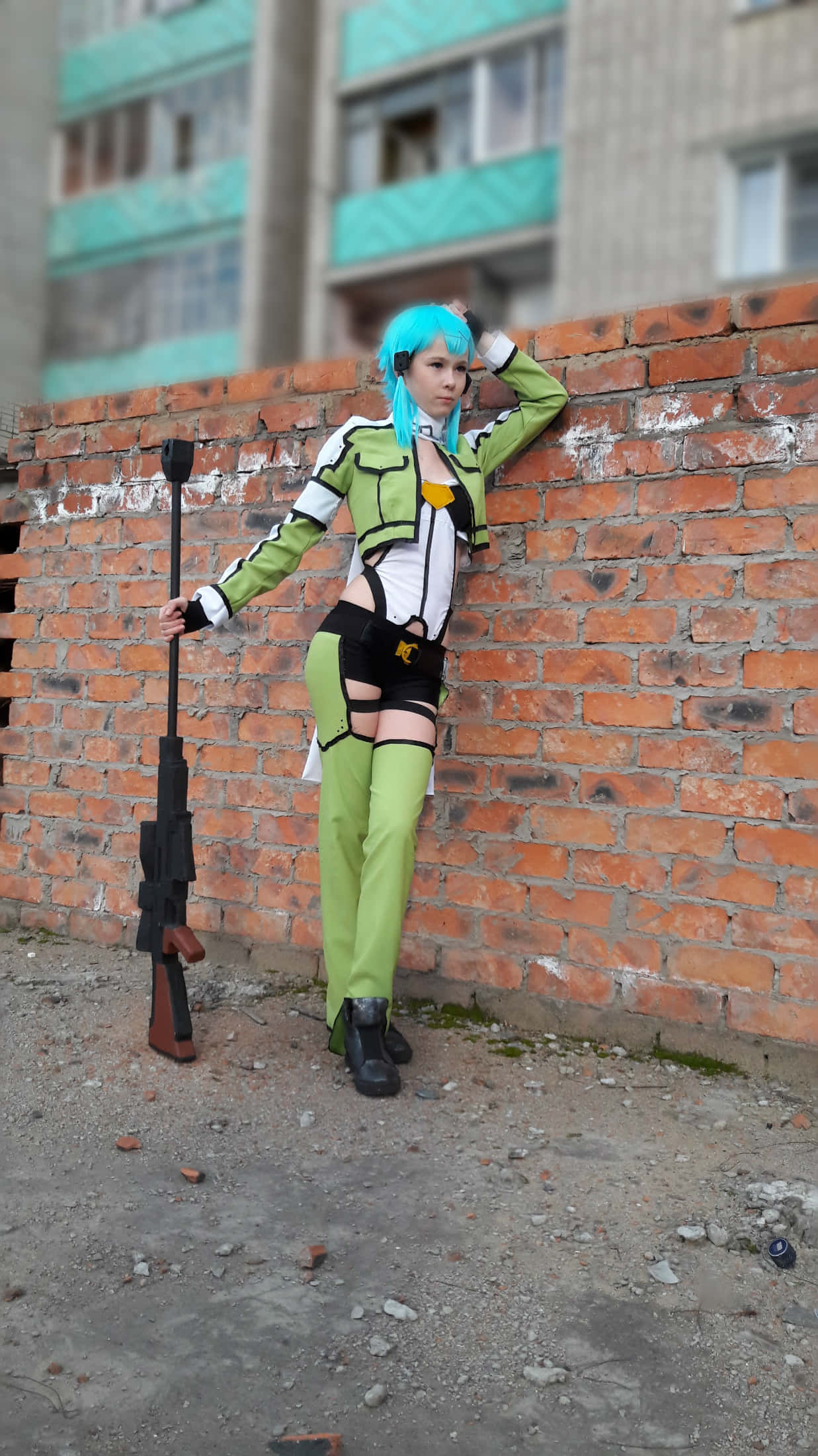 Sword Art Online Cosplay 桌布 壁紙 — 一位擁有亮藍色頭髮和綠色服裝的角色扮演者，在城市環境中靠著磚牆，手持步槍。
