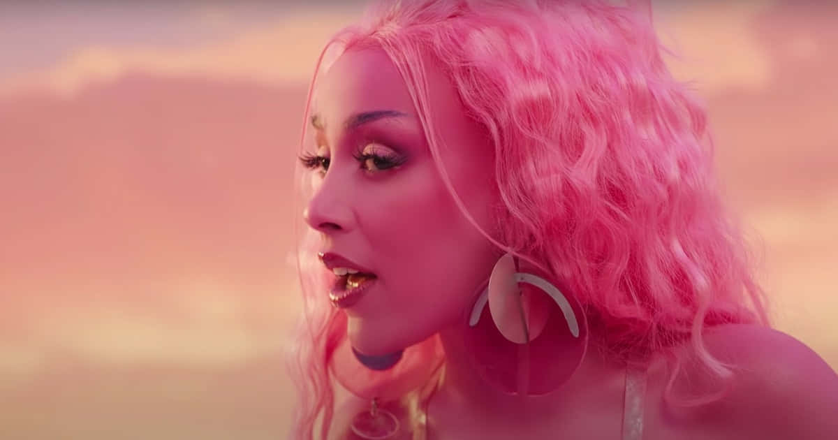 Doja Cat 桌布 壁紙 — 一位擁有充滿活力的粉紅色捲髮和大圈耳環的女性，在柔和的粉紅色日落天空下若有所思地凝視著。
