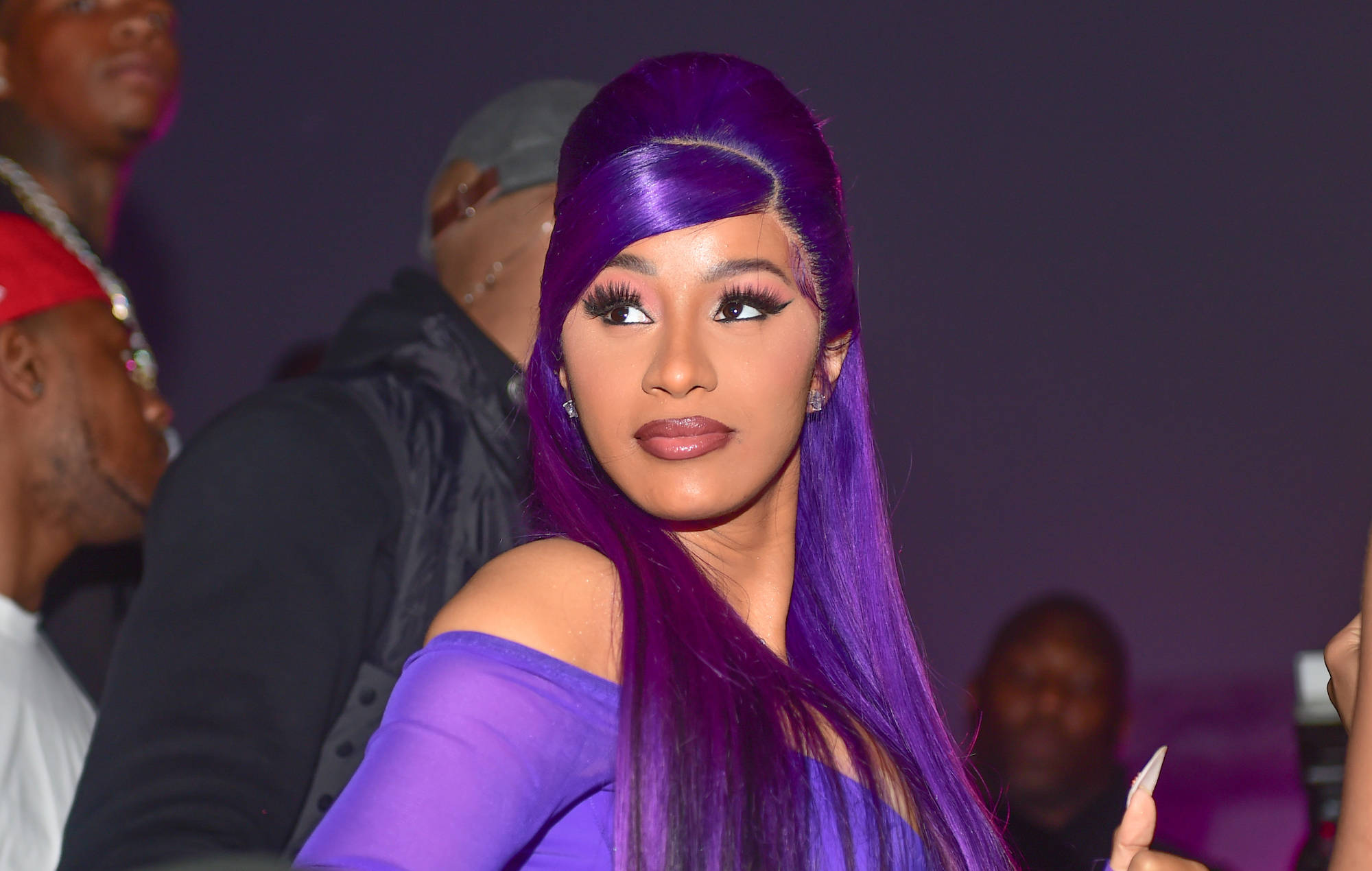 cardi 桌布 壁紙 — 一位擁有充滿活力的紫色頭髮和相配服裝的女性，在燈光昏暗的活動中自信地擺姿勢，背景有模糊的與會者。