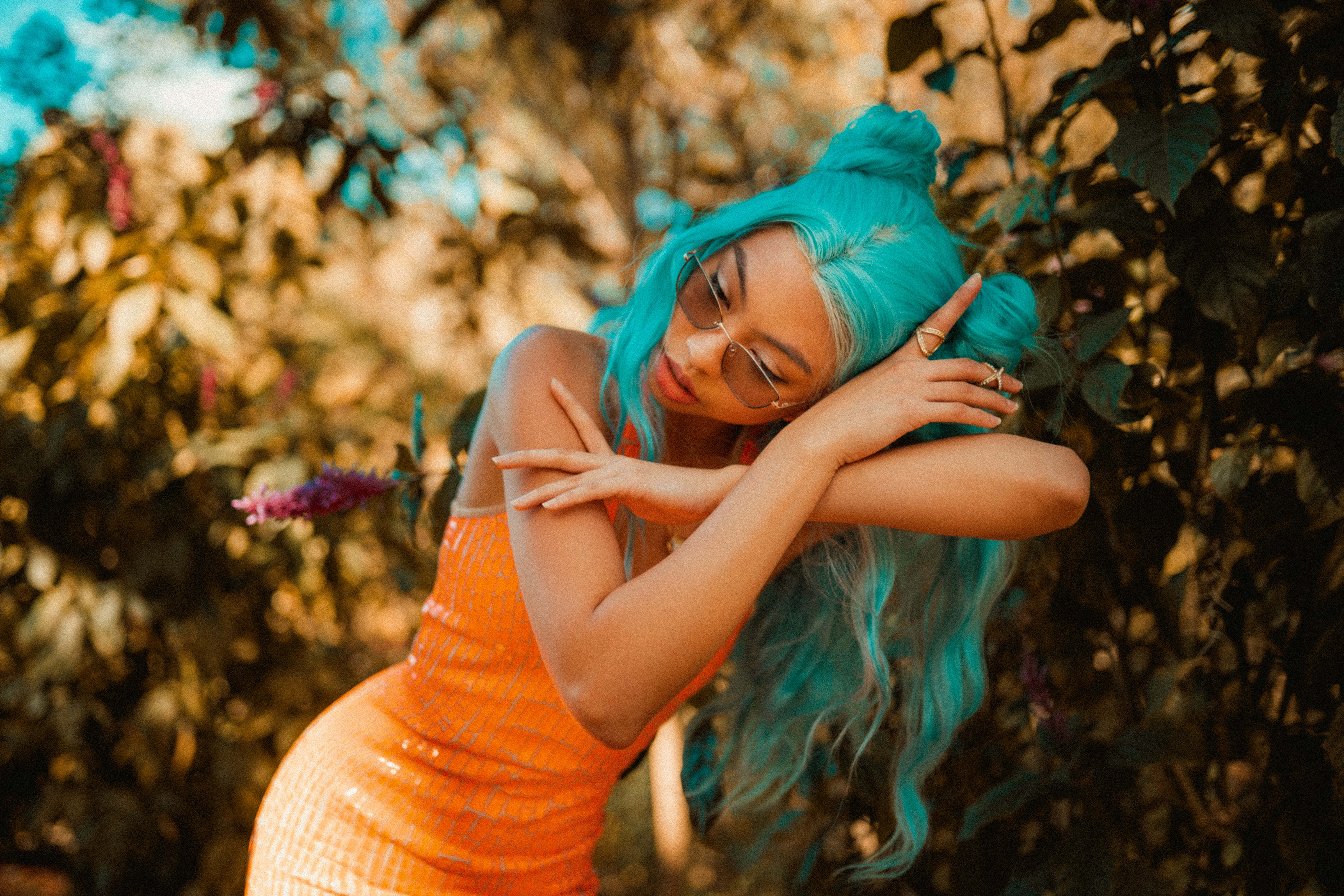 Orange And Teal 桌布 壁紙 — 一位擁有充滿活力的綠松石色頭髮和橙色連衣裙的女性在陽光普照的樹葉中優雅地擺姿勢。