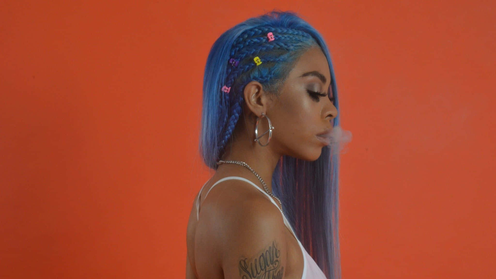 Rico Nasty 桌布 壁紙 — 一位擁有充滿活力的藍色辮子、飾有彩色夾子的女性，在鮮豔的橙色背景前呼出煙霧。