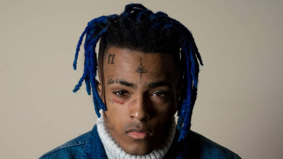 酷酷的xxxtentacion 桌布 壁紙 — 一位擁有充滿活力的藍色辮子和面部刺青的年輕男子，在純米色背景前強烈凝視。