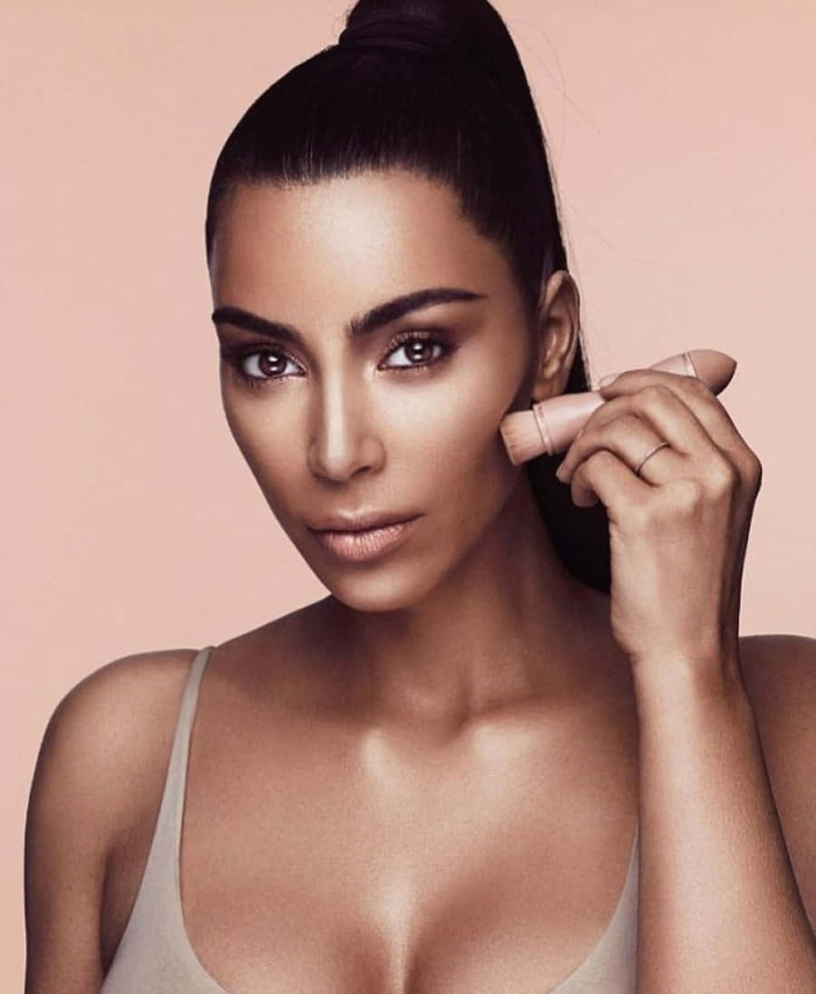 Kim Kardashian 桌布 壁紙 — 一位擁有光滑高馬尾的女性在柔和的蜜桃色背景下將腮紅塗在臉頰上。