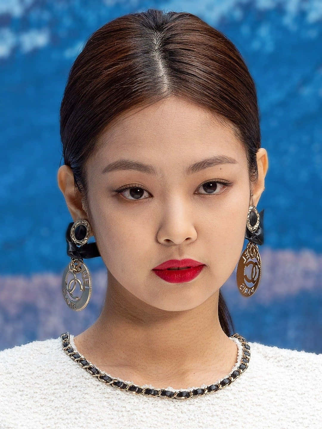 BLACKPINK 桌布 壁紙 — 一位擁有光滑黑髮和大膽紅唇的女性，在帶有紋理的藍色背景前佩戴華麗的香奈兒耳環。