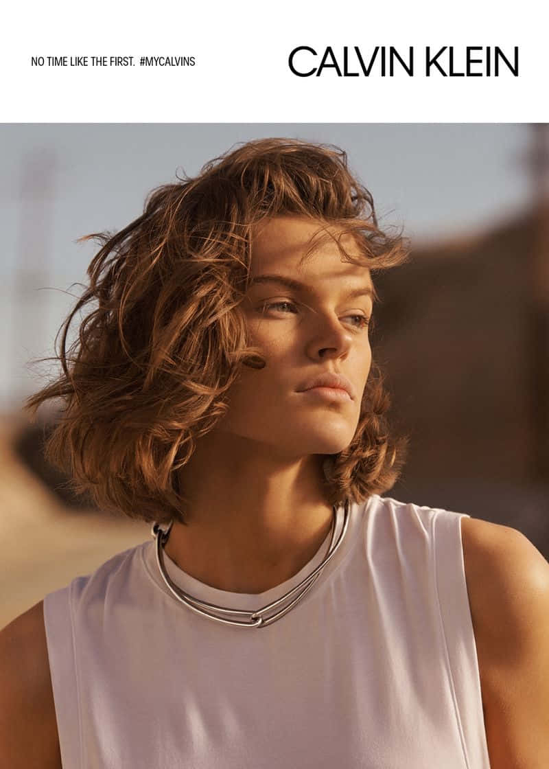 Calvin Klein 桌布 壁紙 — 一位擁有凌亂棕色頭髮的模特兒，在溫暖的沙漠背景下，穿著白色無袖上衣和層疊的銀色項鍊。