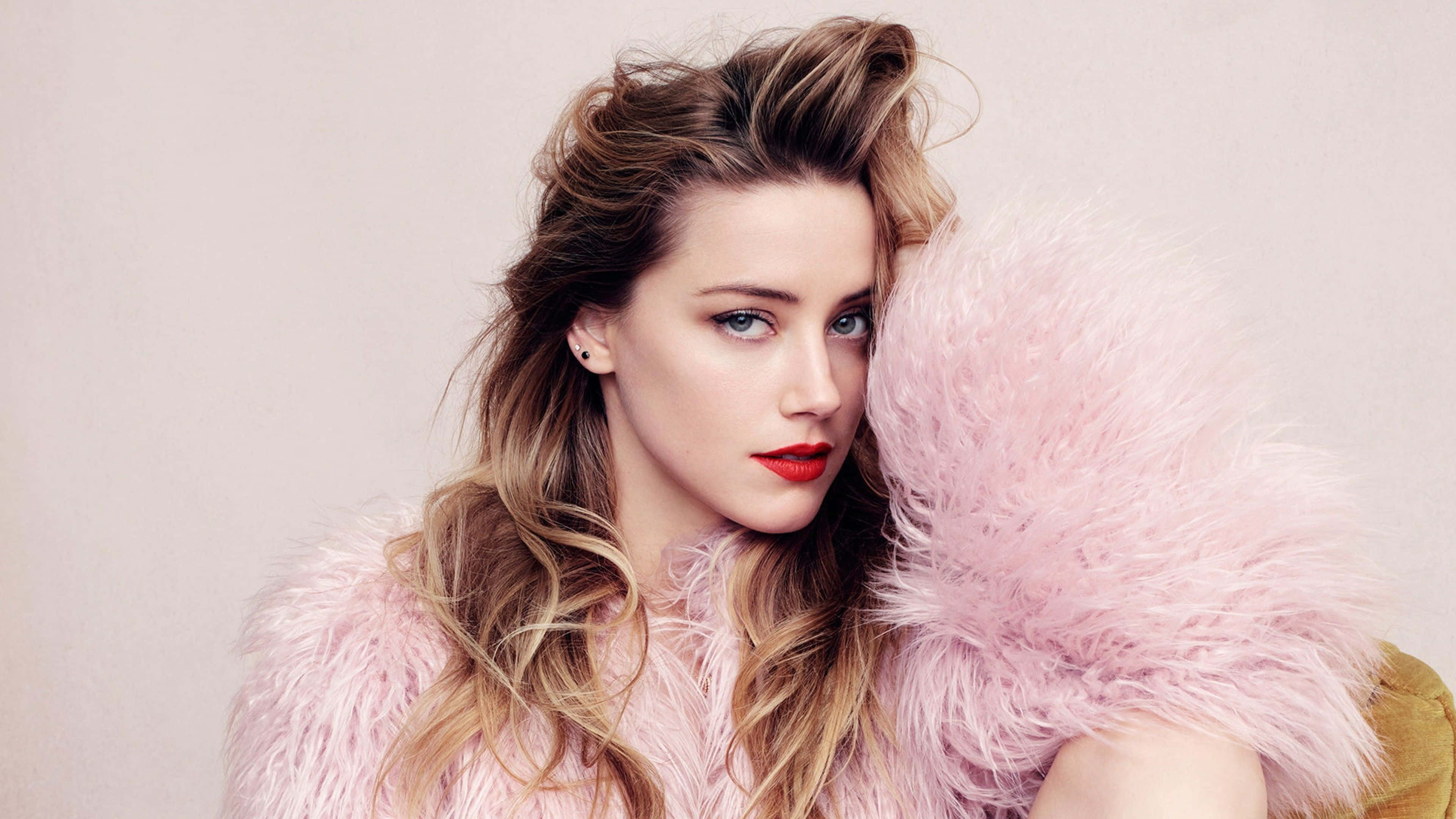 Amber Heard 桌布 壁紙 — 一位擁有凌亂金髮和大膽紅唇的女性，在中性背景前穿著柔軟的粉紅色羽毛外套。