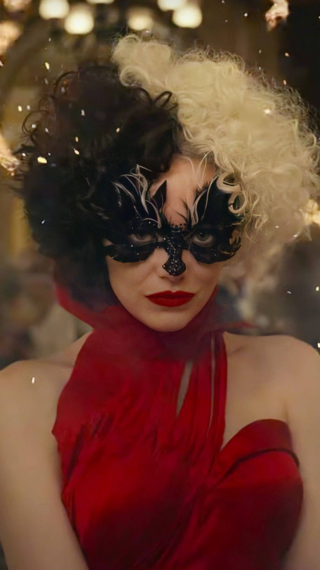 Cruella 2021 桌布 壁紙 — 一位擁有半黑半白頭髮的女性，在溫暖的燈光下戴著華麗的黑色面具和鮮豔的紅色連衣裙。