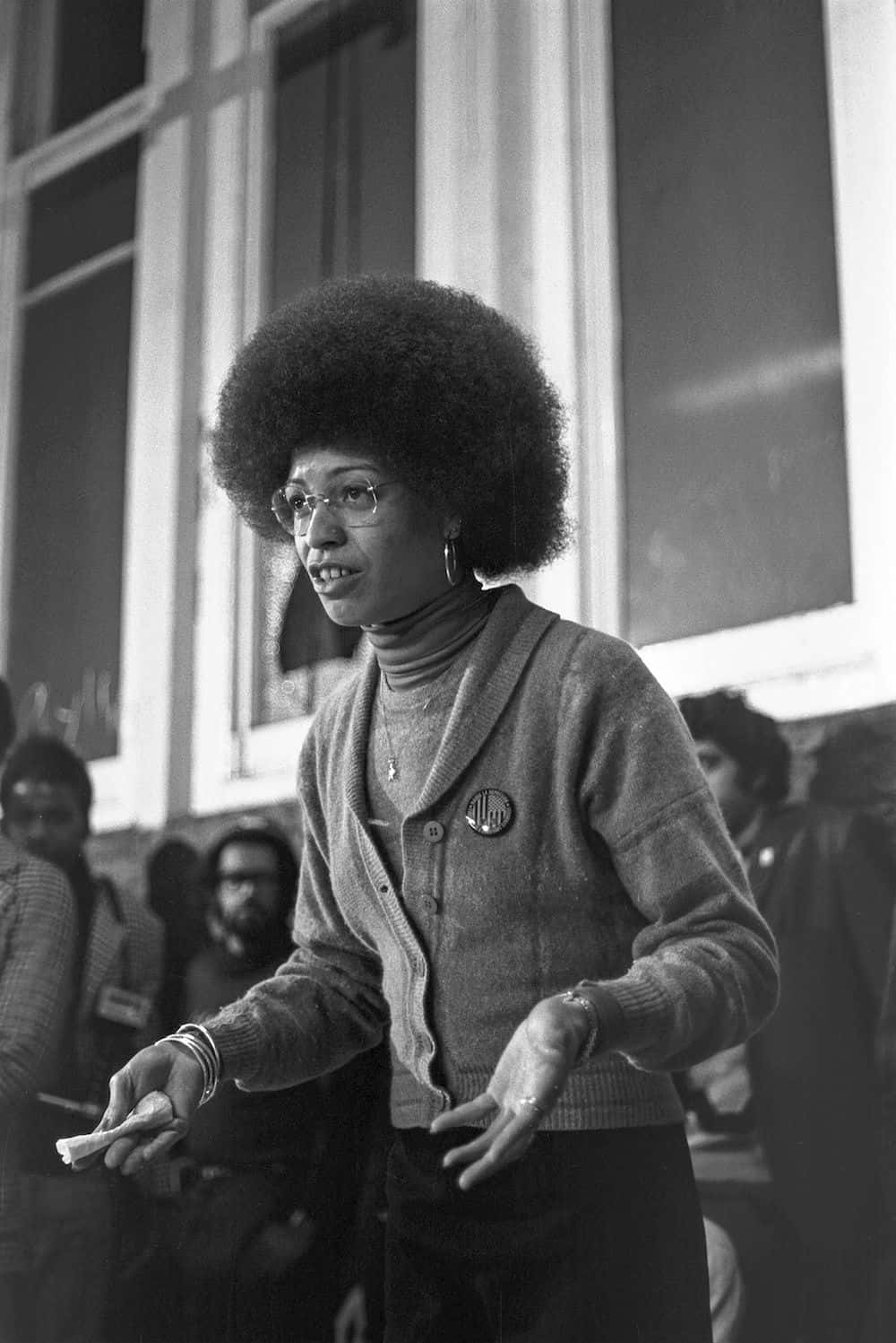 Black Panther Party 桌布 壁紙 — 一位擁有大型爆炸頭和眼鏡的女性在室內活動中說話時做手勢，以黑白拍攝。