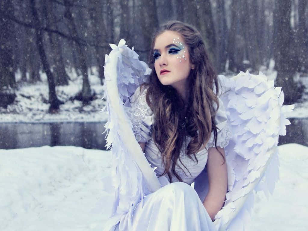 Snow Angel 桌布 壁紙 — 一位擁有大型白色羽毛翅膀的女性，穿著白色連衣裙和閃亮的藍色眼妝，坐在雪地森林中。