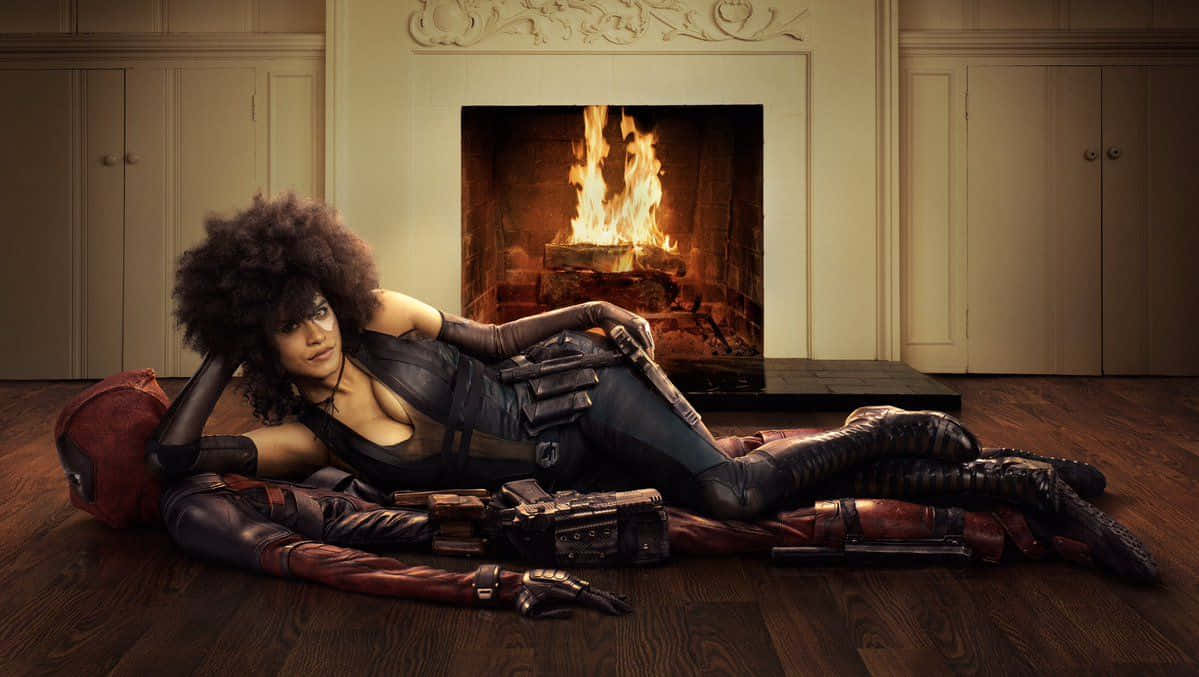 Deadpool And Domino 桌布 壁紙 — 一位擁有大型非洲髮型的女性放鬆地躺在木地板上，旁邊是一個穿著紅色西裝的人物，身後是經典房間中熊熊燃燒的壁爐。