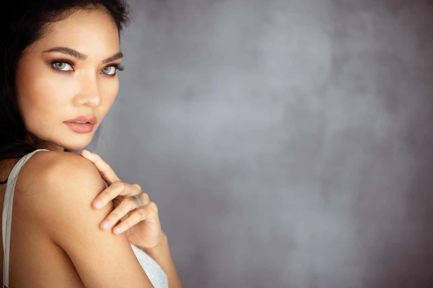 Woman 桌布 壁紙 — 一位擁有引人注目的眼睛和深色頭髮的女性，在紋理灰色背景前優雅地擺姿勢，手放在肩膀上。
