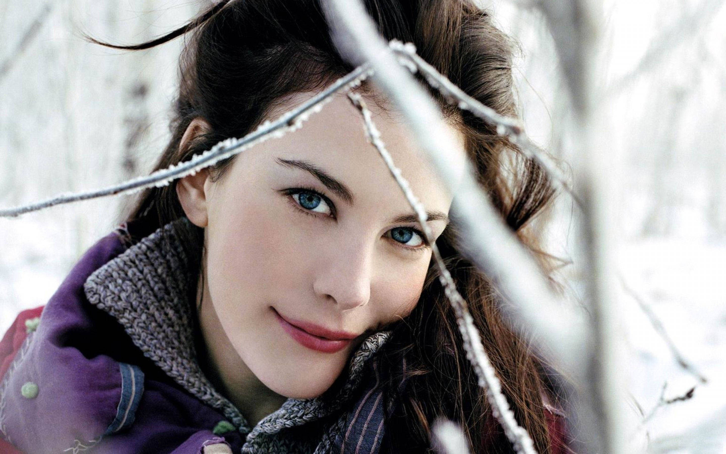 Fashion Model 桌布 壁紙 — 一位擁有引人注目的藍眼睛的女性，在白雪皚皚的冬季森林中，透過結霜的樹枝溫柔地微笑。