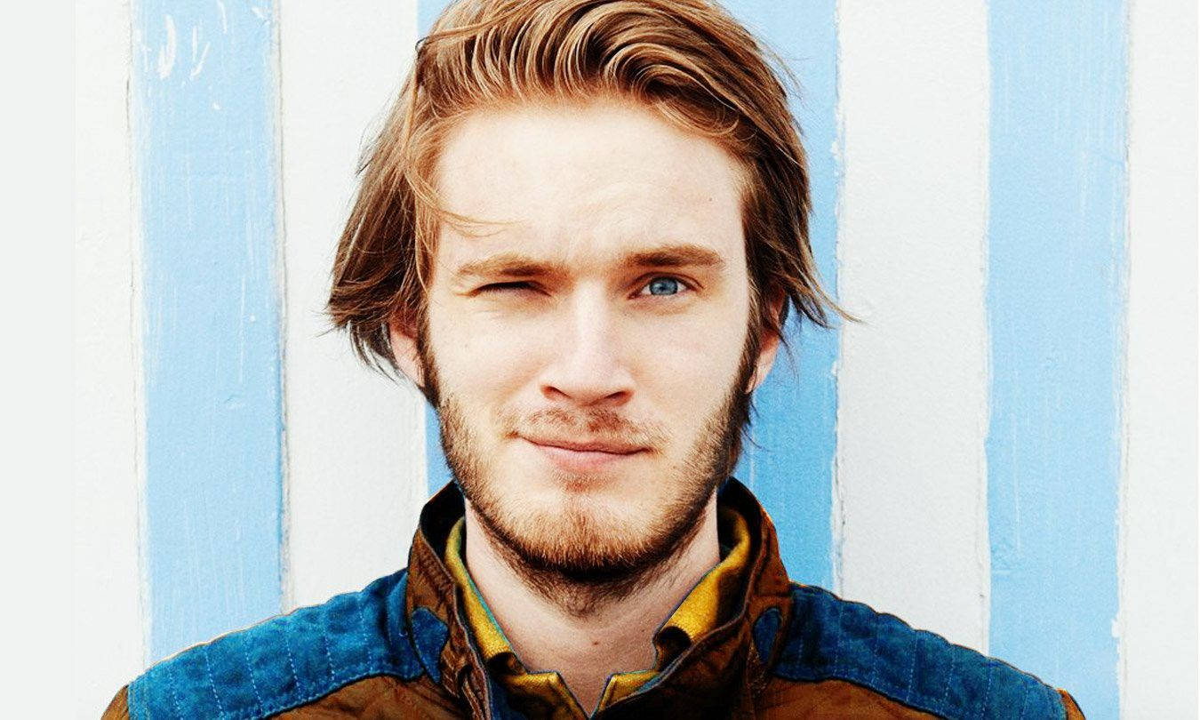 PewDiePie 桌布 壁紙 — 一位擁有引人注目的藍色眼睛和微紅頭髮的年輕男子，在藍白條紋牆前頑皮地眨眼。