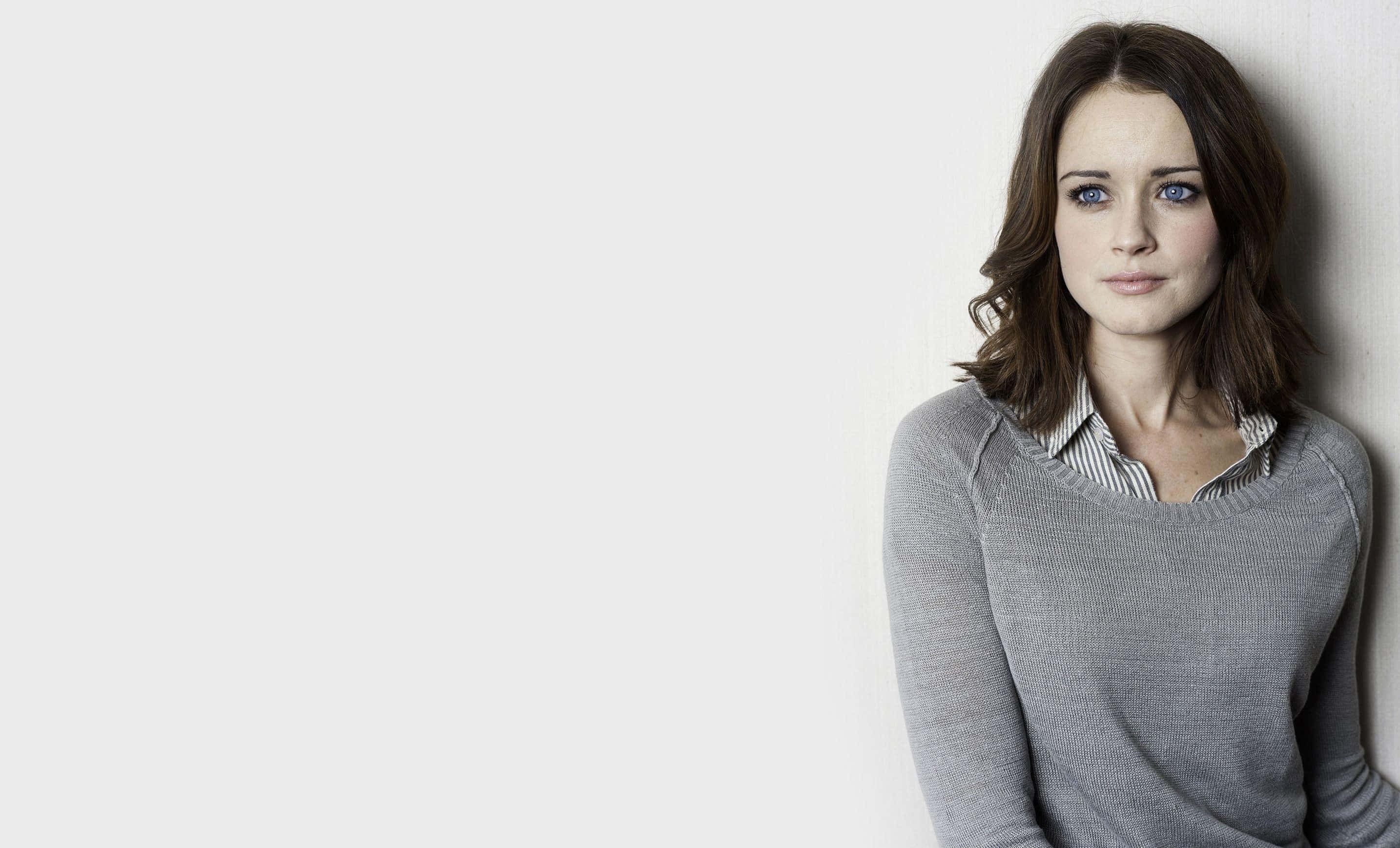 Alexis Bledel 桌布 壁紙 — 一位擁有引人注目的藍色眼睛和棕色頭髮的女性，靠在一面純白色的牆上，穿著灰色毛衣，內搭條紋襯衫。