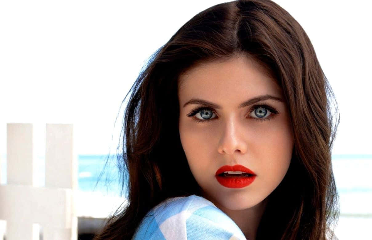 Alexandra Daddario 桌布 壁紙 — 一位擁有引人注目的藍色眼睛和紅色嘴唇的女性，在明亮的沙灘背景下強烈凝視。