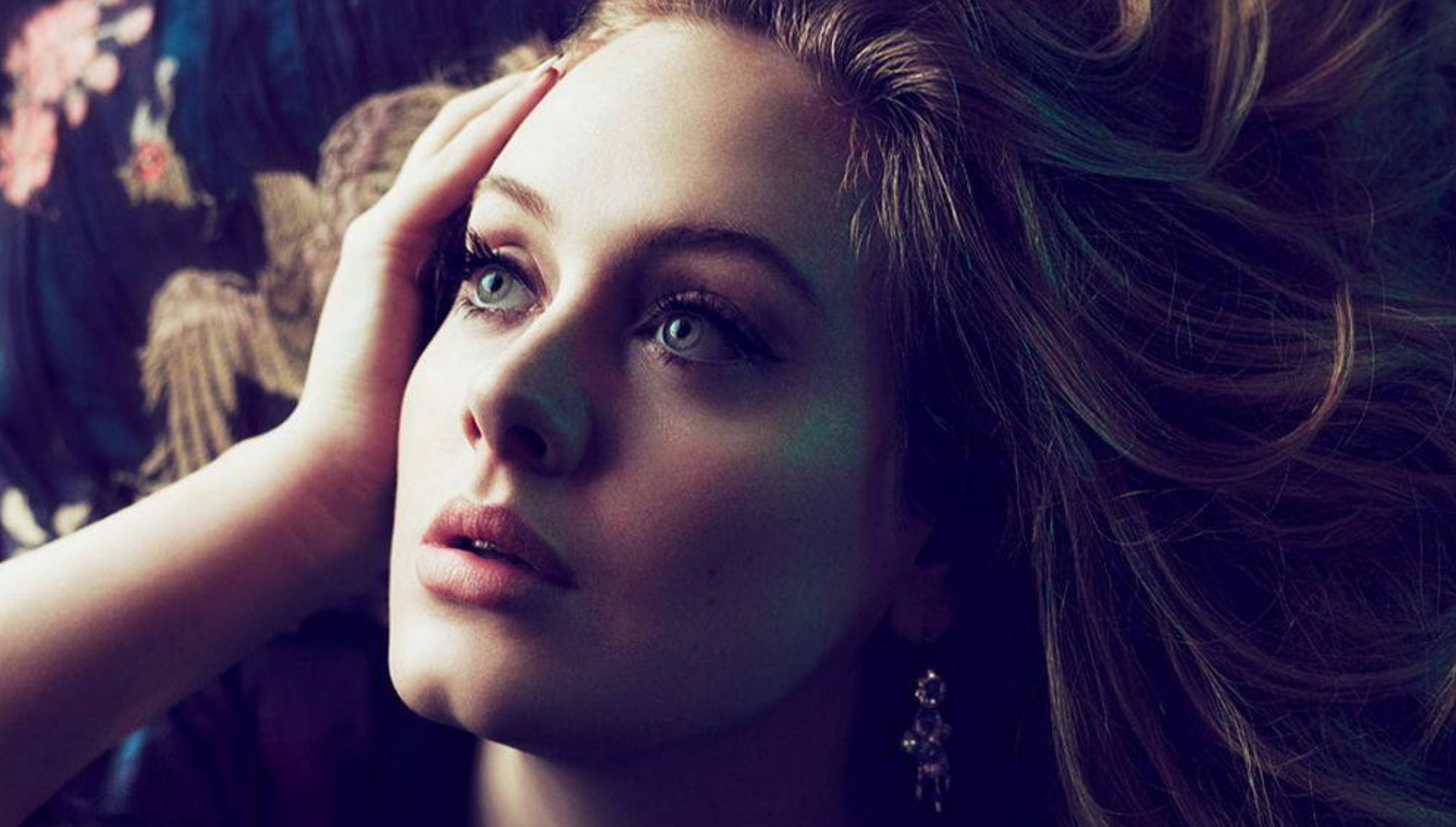 Adele 桌布 壁紙 — 一位擁有引人注目的藍色眼睛和飄逸長髮的女性，在花卉圖案背景前將手放在額頭上。