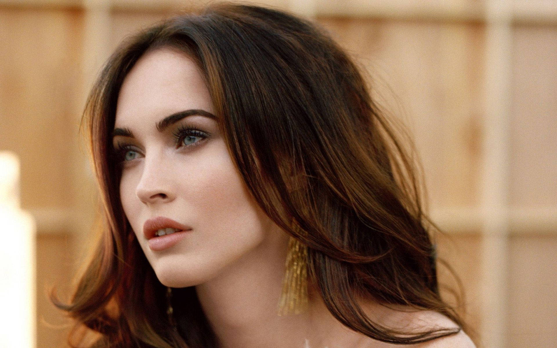 Megan Fox HD 桌布 壁紙 — 一位擁有引人注目的藍色眼睛和飄逸棕色頭髮的女性特寫肖像，戴著金色流蘇耳環。