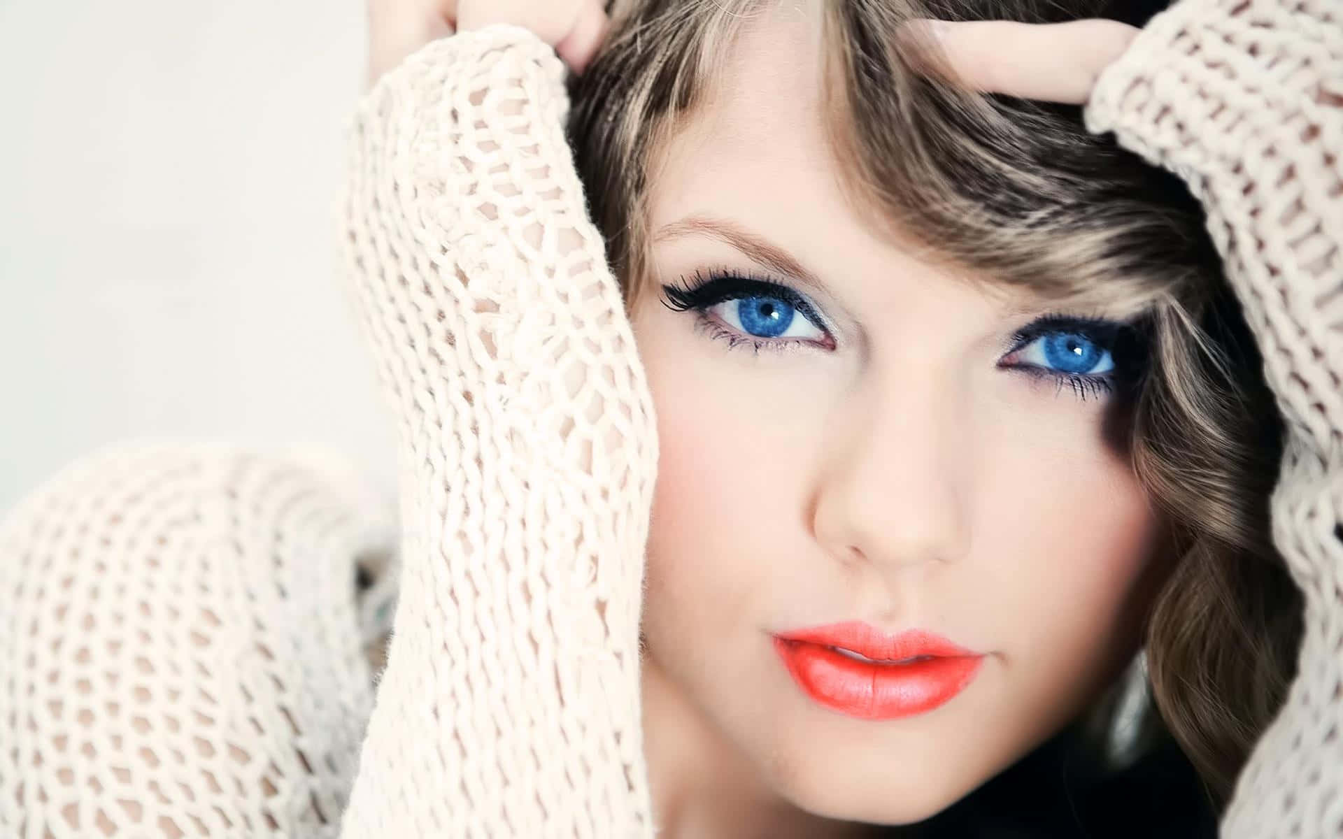 Taylor Swift Lover 桌布 壁紙 — 一位擁有引人注目藍眼睛和紅唇的女性，從奶油色針織毛衣中探出頭來的特寫肖像。