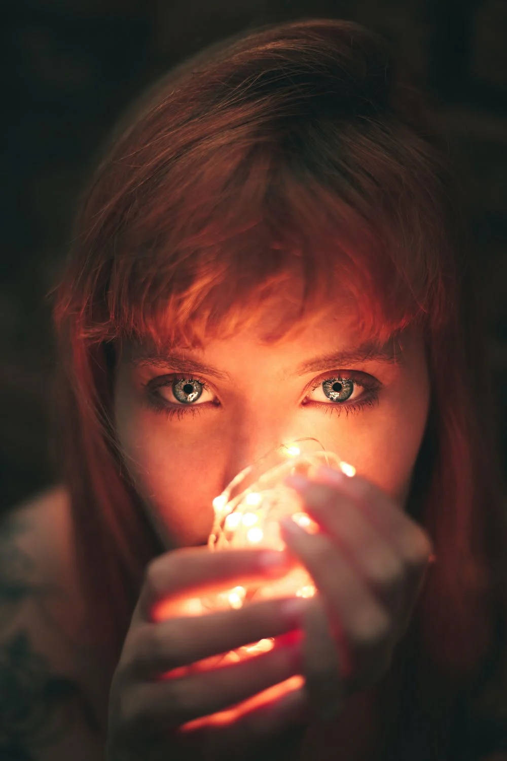 手機 桌布 壁紙 — 一位擁有引人注目藍眼睛的年輕女性，在溫暖昏暗的燈光下，將發光的燈串靠近臉龐，凝視著。