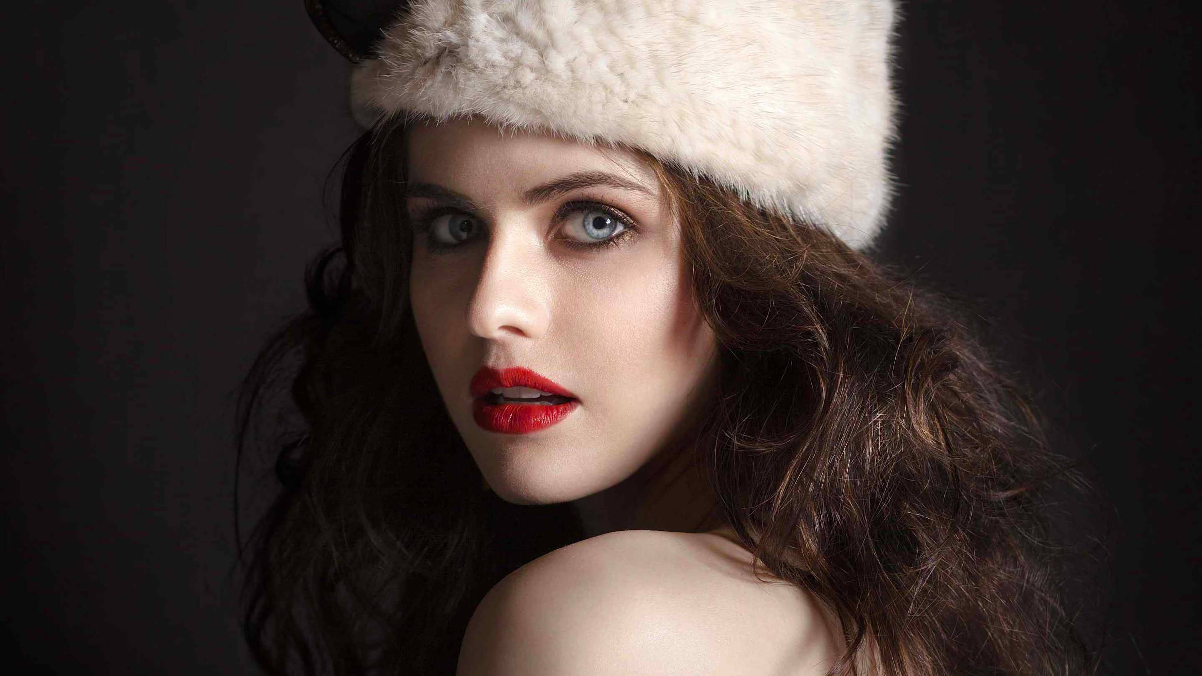 Alexandra Daddario 4K 桌布 壁紙 — 一位擁有引人注目藍色眼睛和紅色嘴唇的女性，在深色背景下戴著一頂蓬鬆的白色帽子。