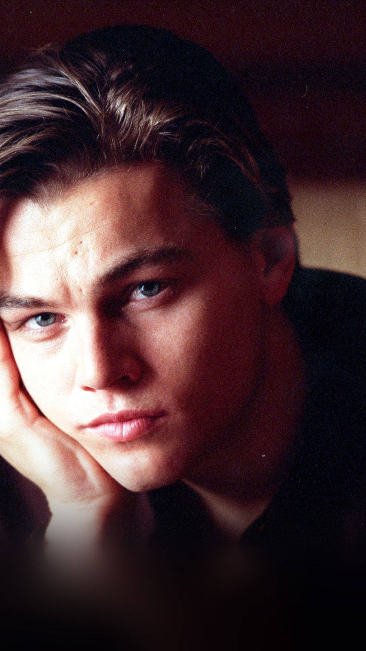 Dicaprio 桌布 壁紙 — 一位擁有強烈藍眼睛的年輕男子在昏暗燈光下，以溫暖色調的肖像中將頭靠在手上。