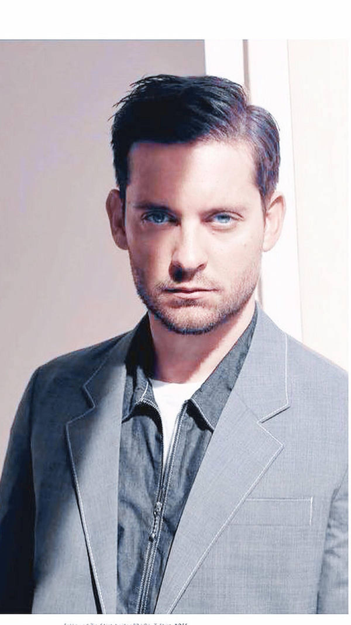 Tobey Maguire 桌布 壁紙 — 一位擁有強烈藍色眼睛和時尚深色頭髮的男子，在純色淺色背景前，穿著灰色西裝外套在深色拉鍊夾克外面。