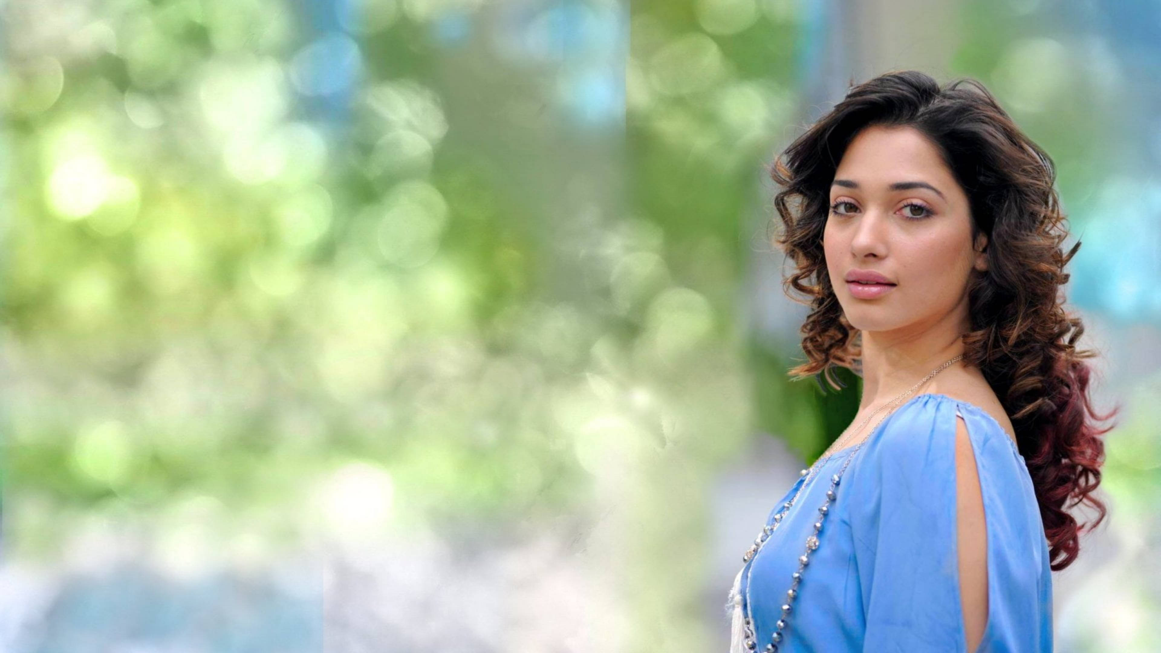 tamannaah bhatia 桌布 壁紙 — 一位擁有捲曲棕色頭髮的女性穿著藍色上衣，在柔和的綠色散景背景下回頭看。