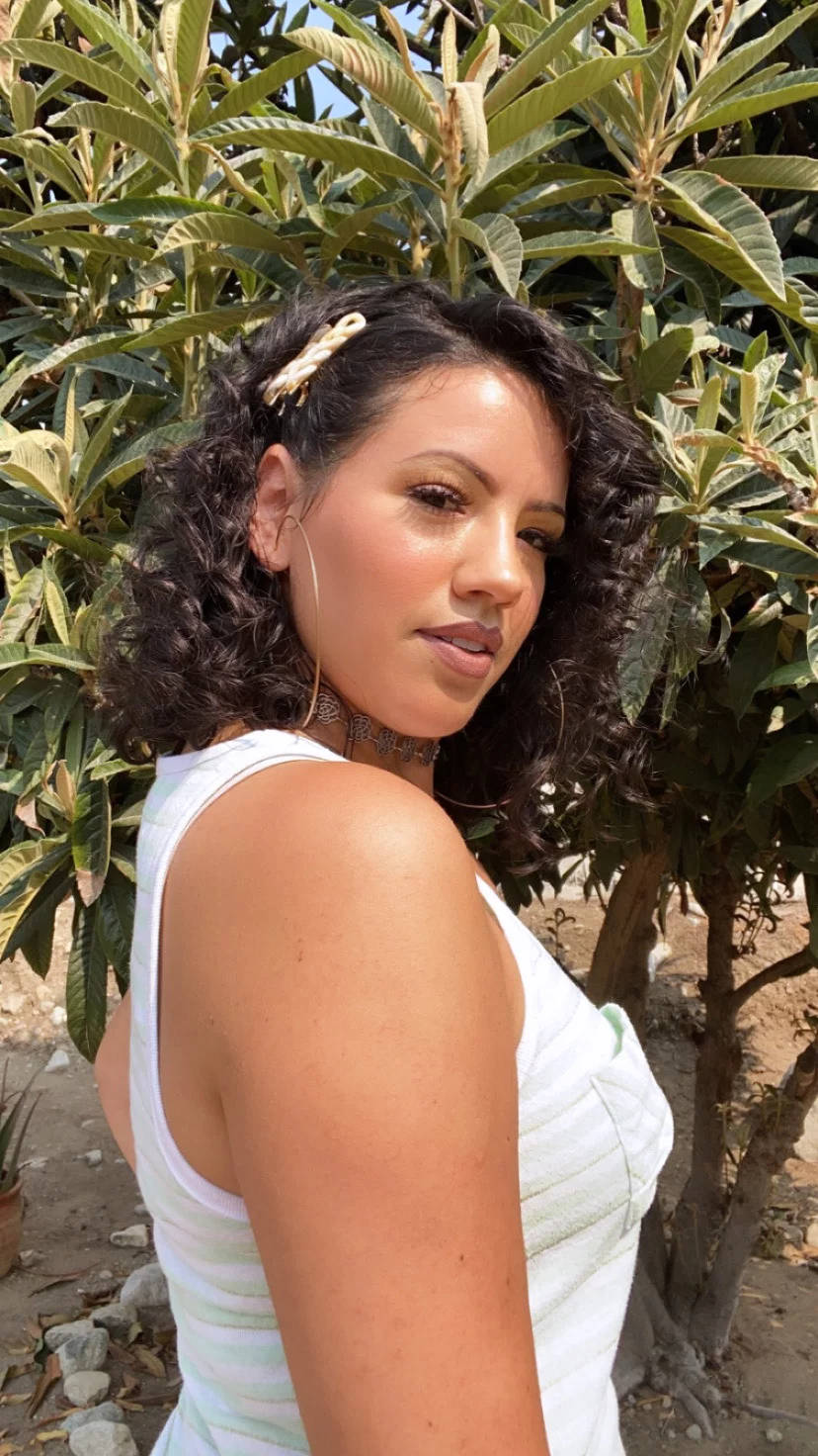 Latina Girl 桌布 壁紙 — 一位擁有捲曲深色頭髮、身穿白色上衣的女性，在明亮的日光下，於茂密的綠色植物旁戶外擺姿勢。