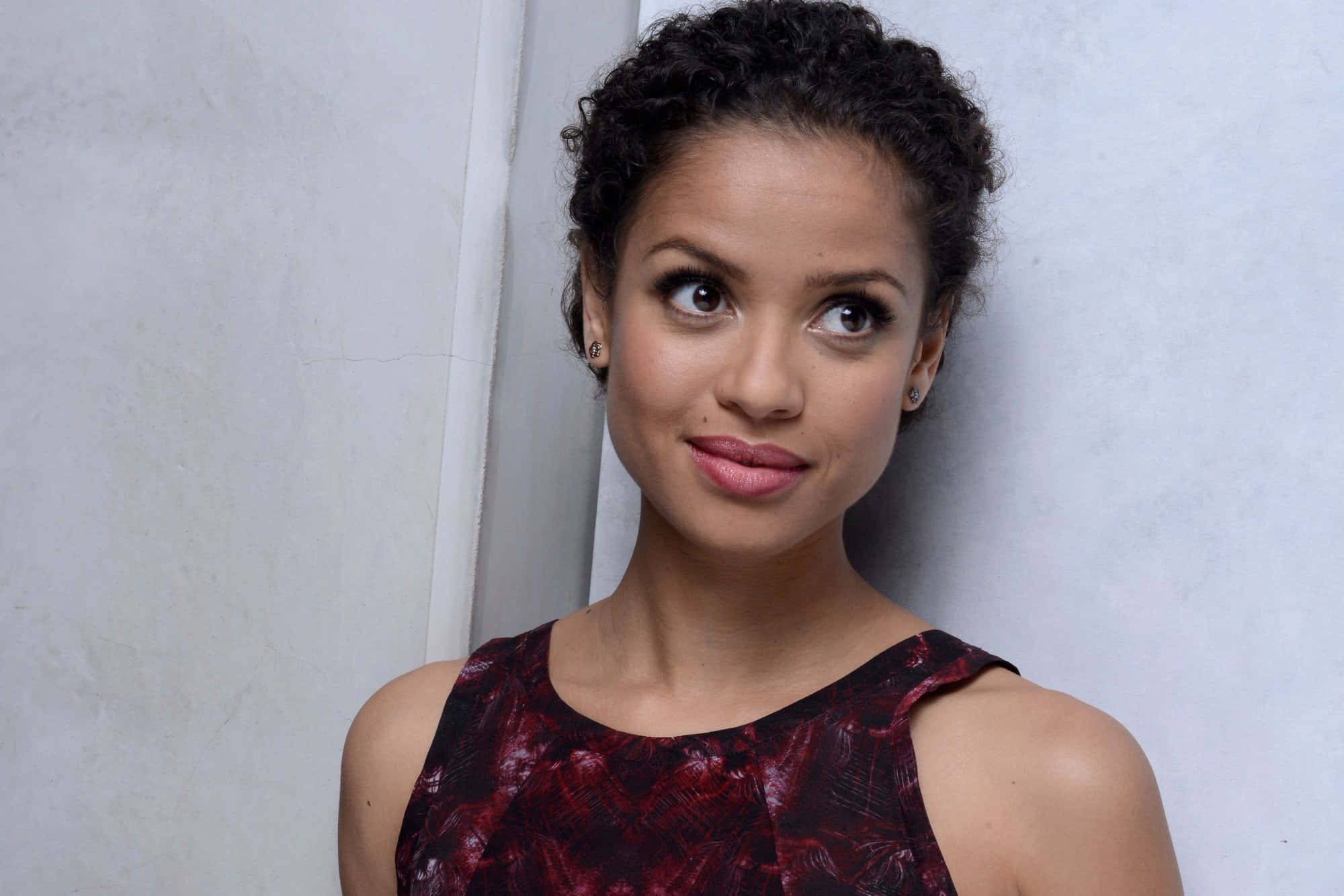 Gugu Mbatha-raw 桌布 壁紙 — 一位擁有捲曲深色頭髮、身穿紅色圖案上衣的女性，帶著俏皮的側視靠在一面素牆上。