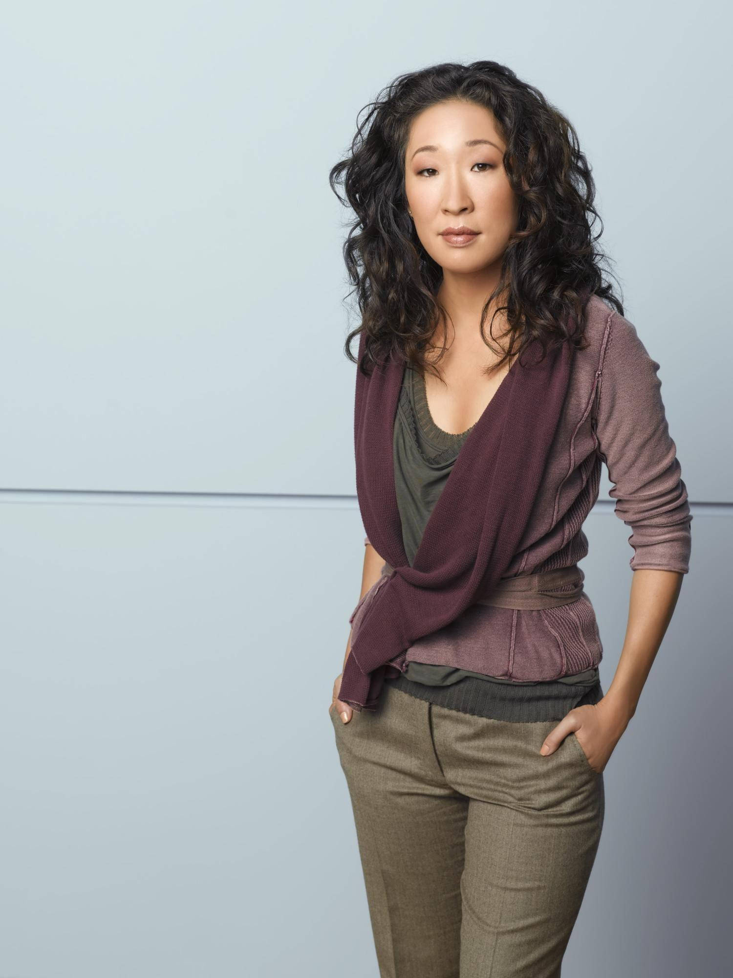 greys anatomy 桌布 壁紙 — 一位擁有捲曲深色頭髮的優雅女性，站在純色淺灰色工作室背景前，穿著多層次的大地色系服裝。