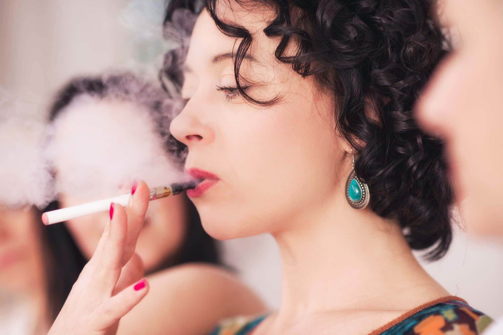 women 桌布 壁紙 — 一位擁有捲曲深色頭髮的女性，戴著綠松石耳環和紅色指甲油，呼出電子煙的蒸氣。