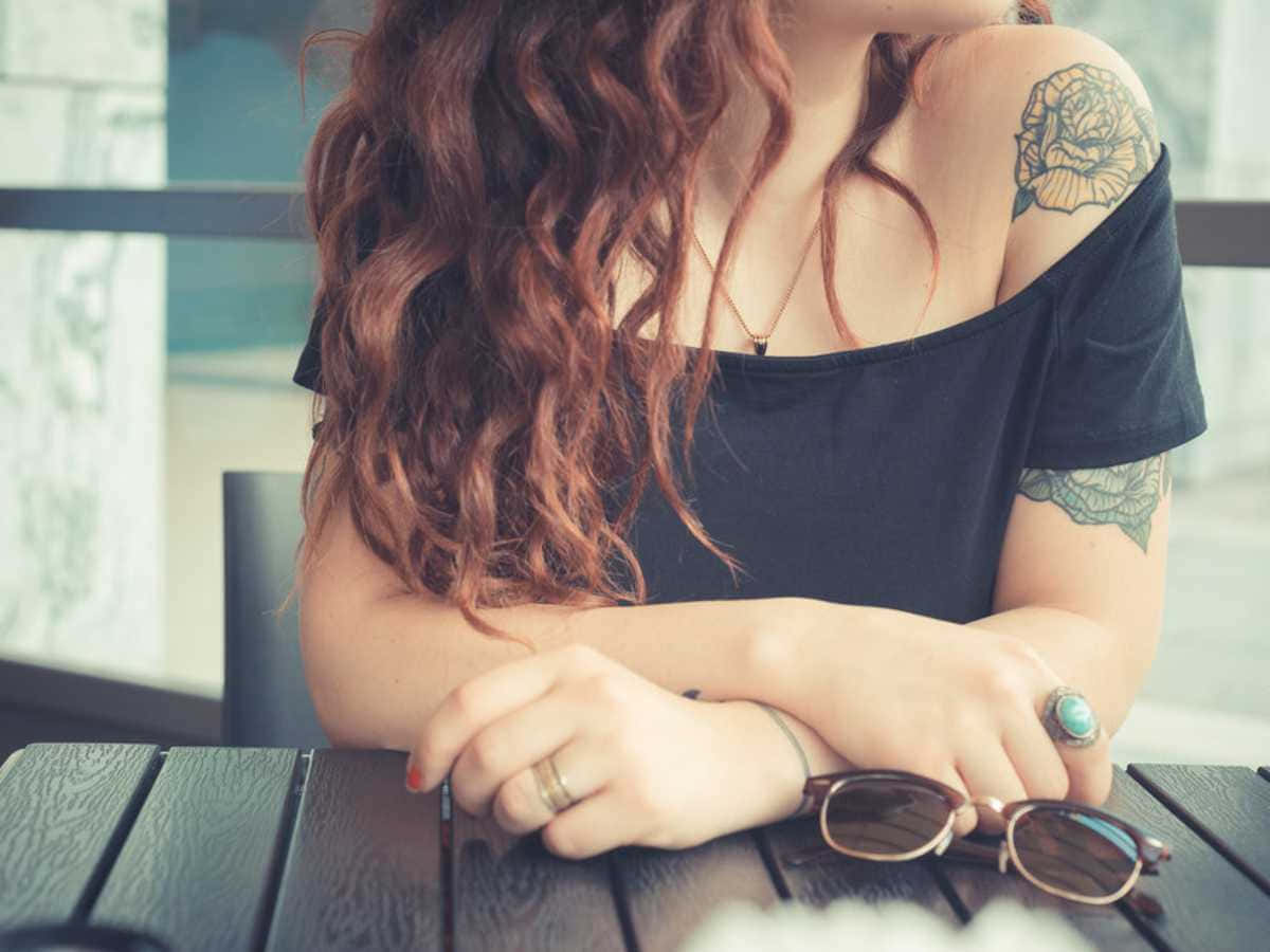 Tattoo Girl 桌布 壁紙 — 一位擁有捲曲赤褐色頭髮和玫瑰刺青的女士，身穿黑色露肩上衣，將手臂擱在木桌上。