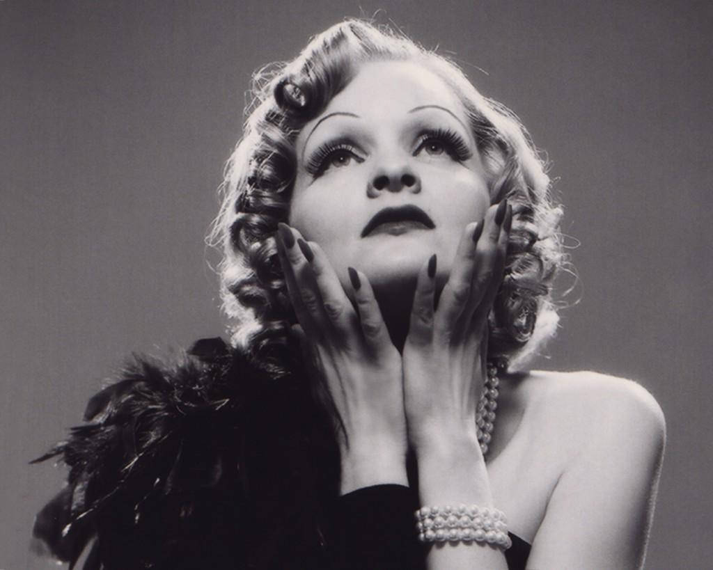 Marlene Dietrich 桌布 壁紙 — 一位擁有捲曲金髮和深色指甲的魅力女性，在經典黑白肖像中雙手托腮向上凝視。