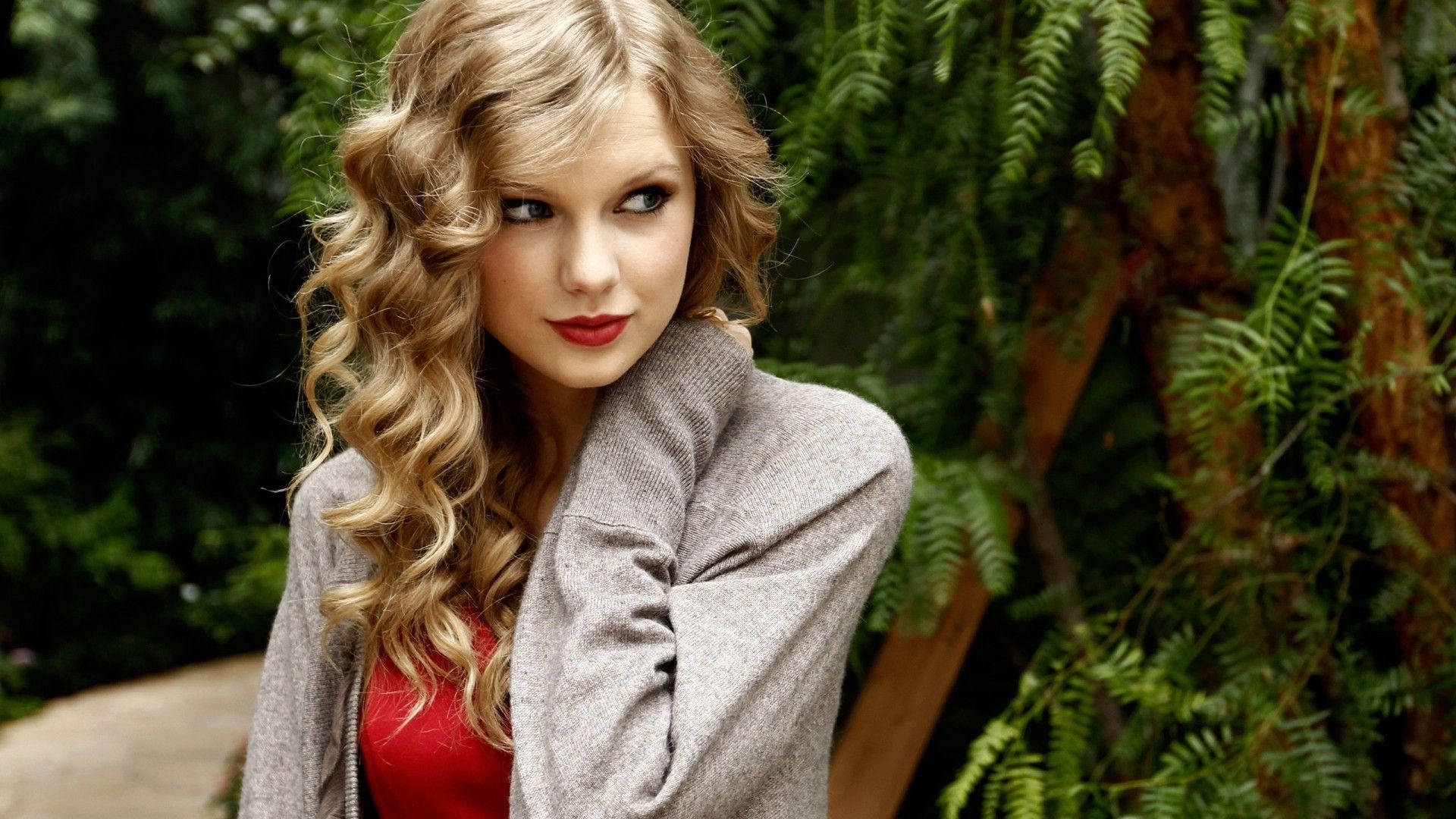 Taylor Swift 桌布 壁紙 — 一位擁有捲曲金髮和紅唇的女性，穿著灰色開襟衫，在茂密的綠色蕨類植物背景下擺姿勢。