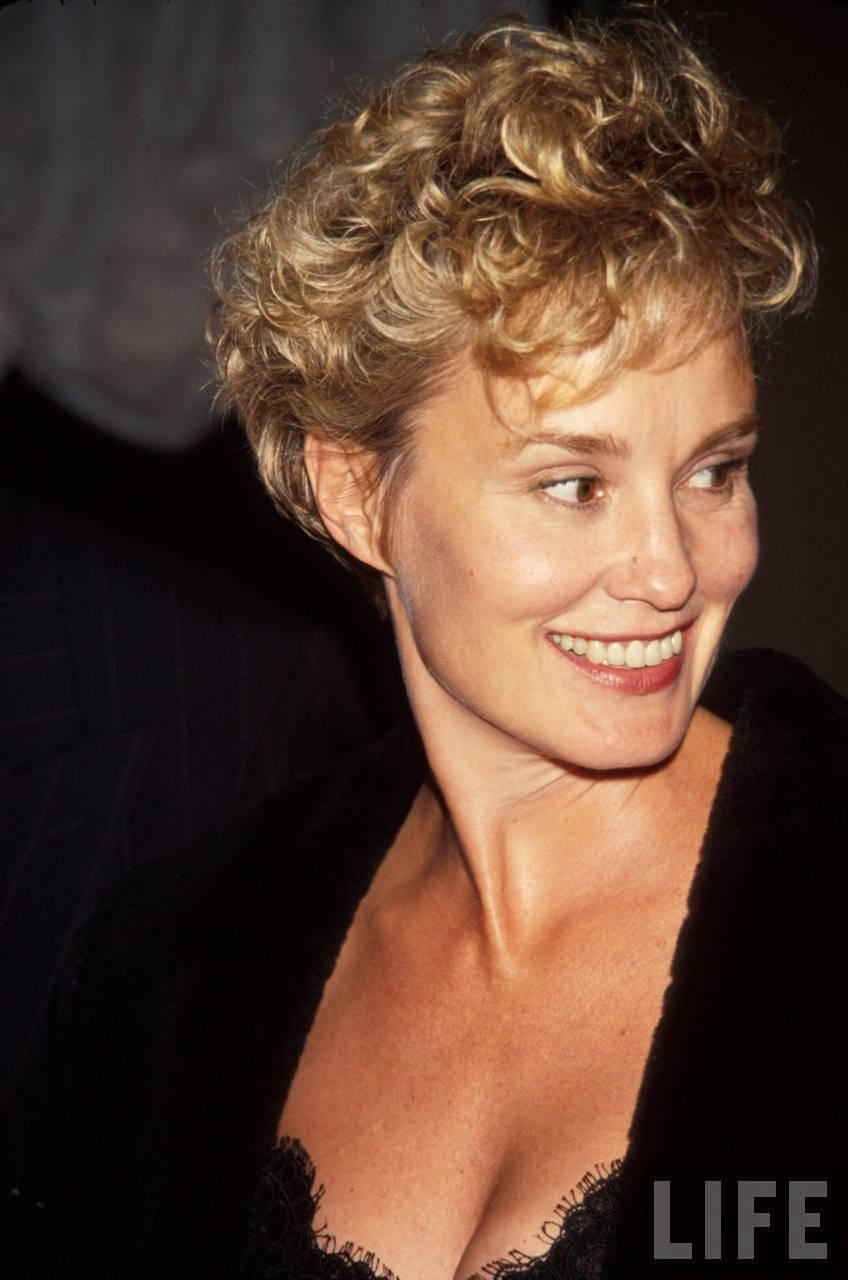 Jessica Lange 桌布 壁紙 — 一位擁有捲曲金髮的女性，穿著黑色服裝，在深色背景下溫暖地微笑。
