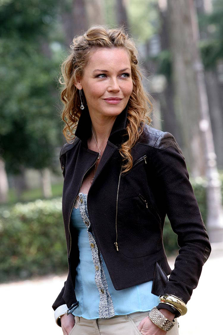 Actress HD 桌布 壁紙 — 一位擁有捲曲金髮的女性，身穿黑色外套和淺藍色上衣，站在陽光明媚的公園裡。