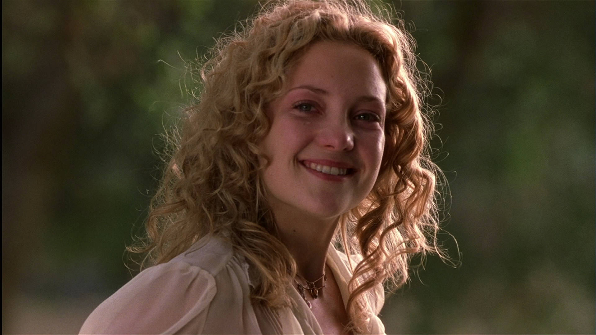 Kate Hudson 桌布 壁紙 — 一位擁有捲曲金髮的女性在柔和自然光線下，對著模糊的綠色背景溫暖地微笑。
