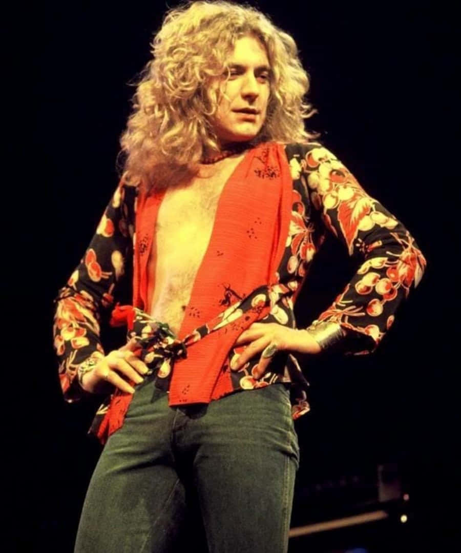 robert plant 桌布 壁紙 — 一位擁有捲曲金髮的搖滾明星在戲劇性的舞台燈光下，穿著花卉圖案外套和紅色圍巾，自信地在舞台上擺姿勢。