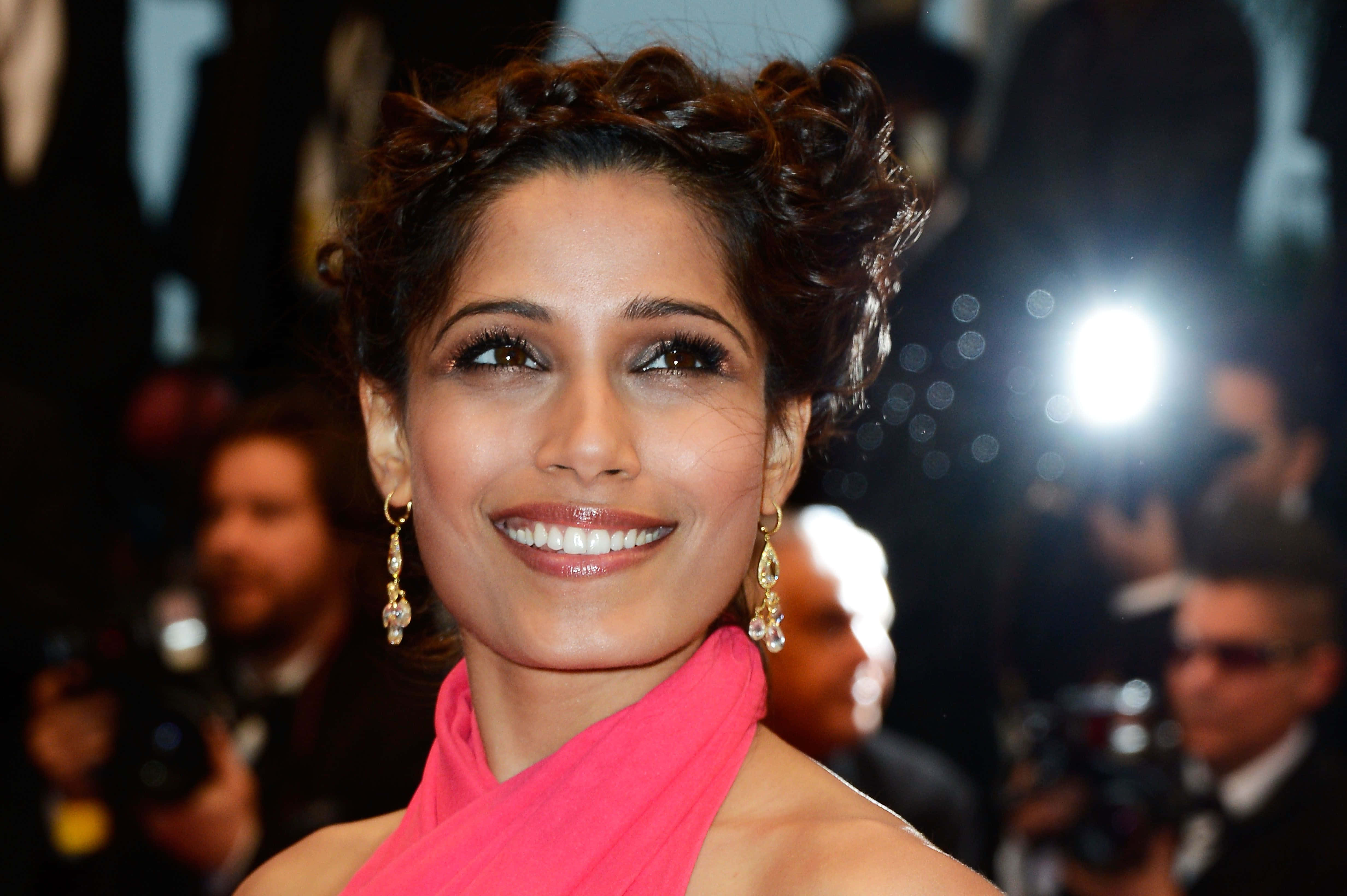 Freida Pinto 桌布 壁紙 — 一位擁有捲髮、戴著粉紅色圍巾的耀眼女性，在背景閃光燈閃爍的華麗活動中燦爛微笑。