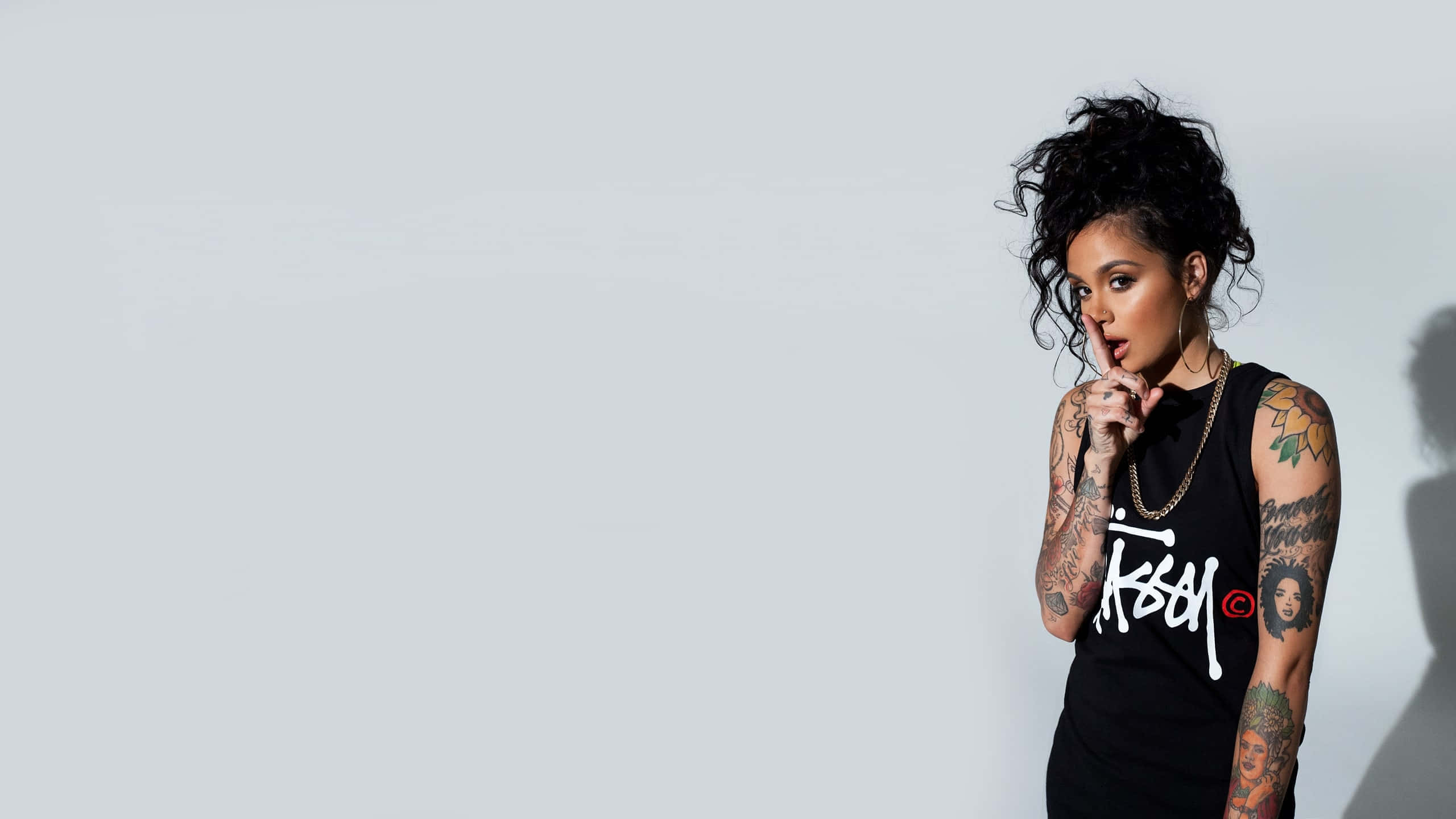 kehlani 桌布 壁紙 — 一位擁有捲髮和刺青的女性，穿著黑色背心，自信地靠在純淺灰色牆壁上擺姿勢。