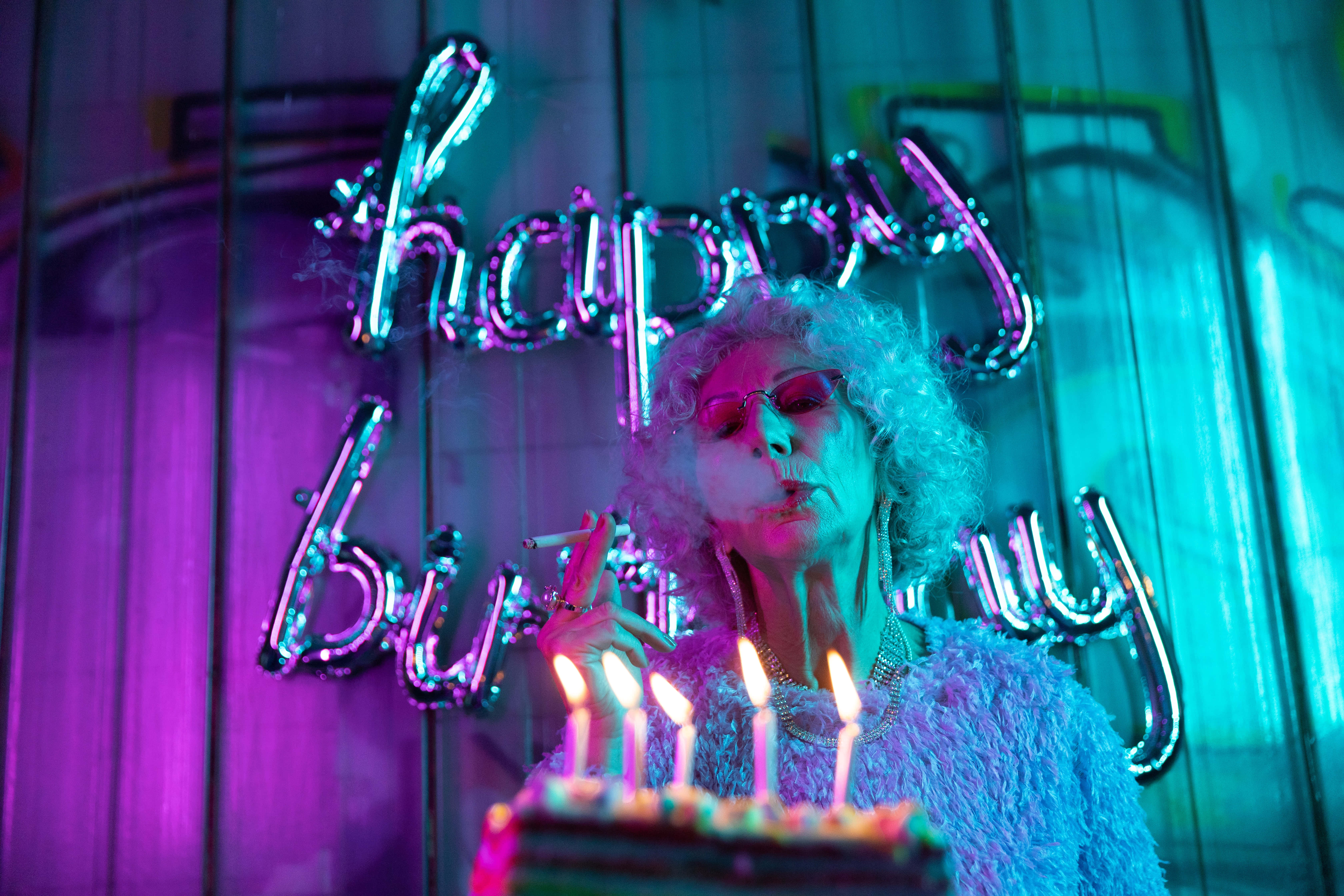 Cute Birthday 桌布 壁紙 — 一位擁有捲髮和太陽眼鏡的女性，在發光的 'happy birthday' 霓虹燈牌下吹熄生日蛋糕上的蠟燭。