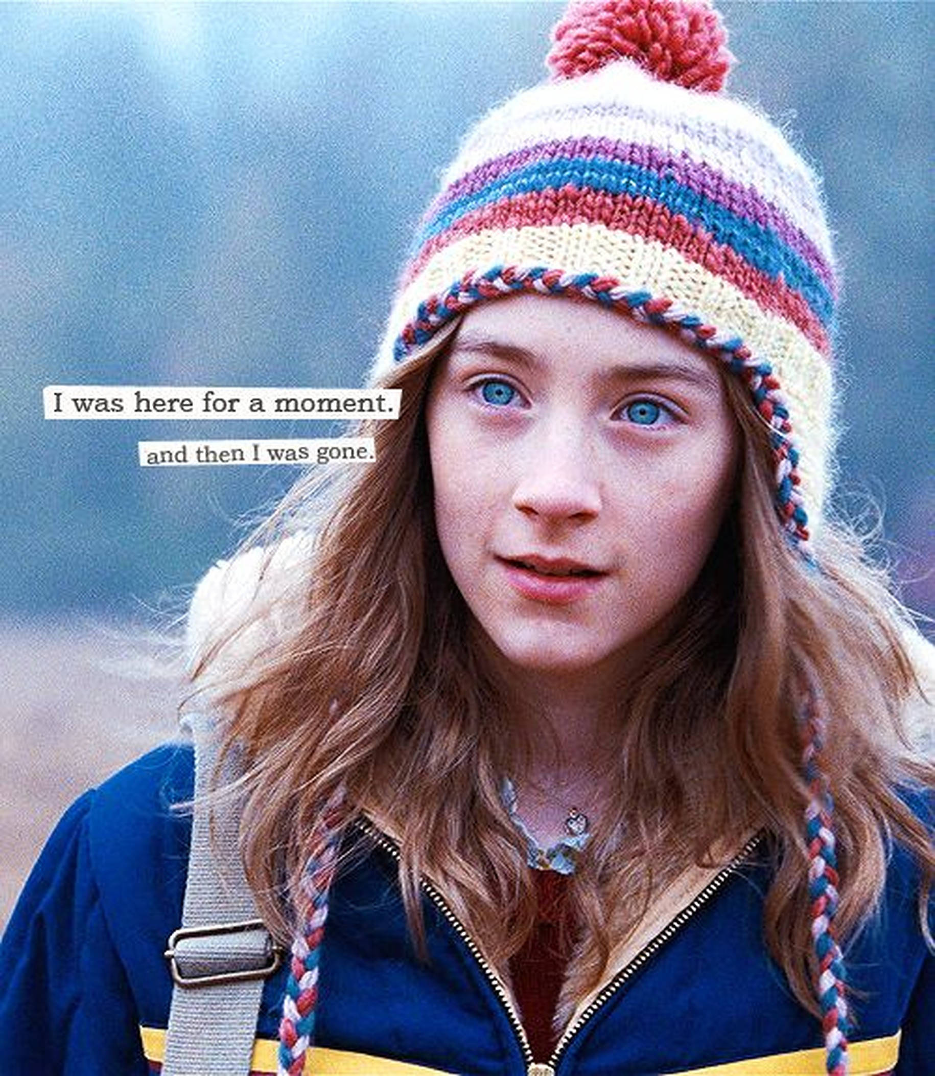 Saoirse Ronan 桌布 壁紙 — 一位擁有明亮藍眼睛的年輕女孩，戴著一頂帶有紅色絨球的彩色條紋針織帽，背景是模糊的戶外景色。