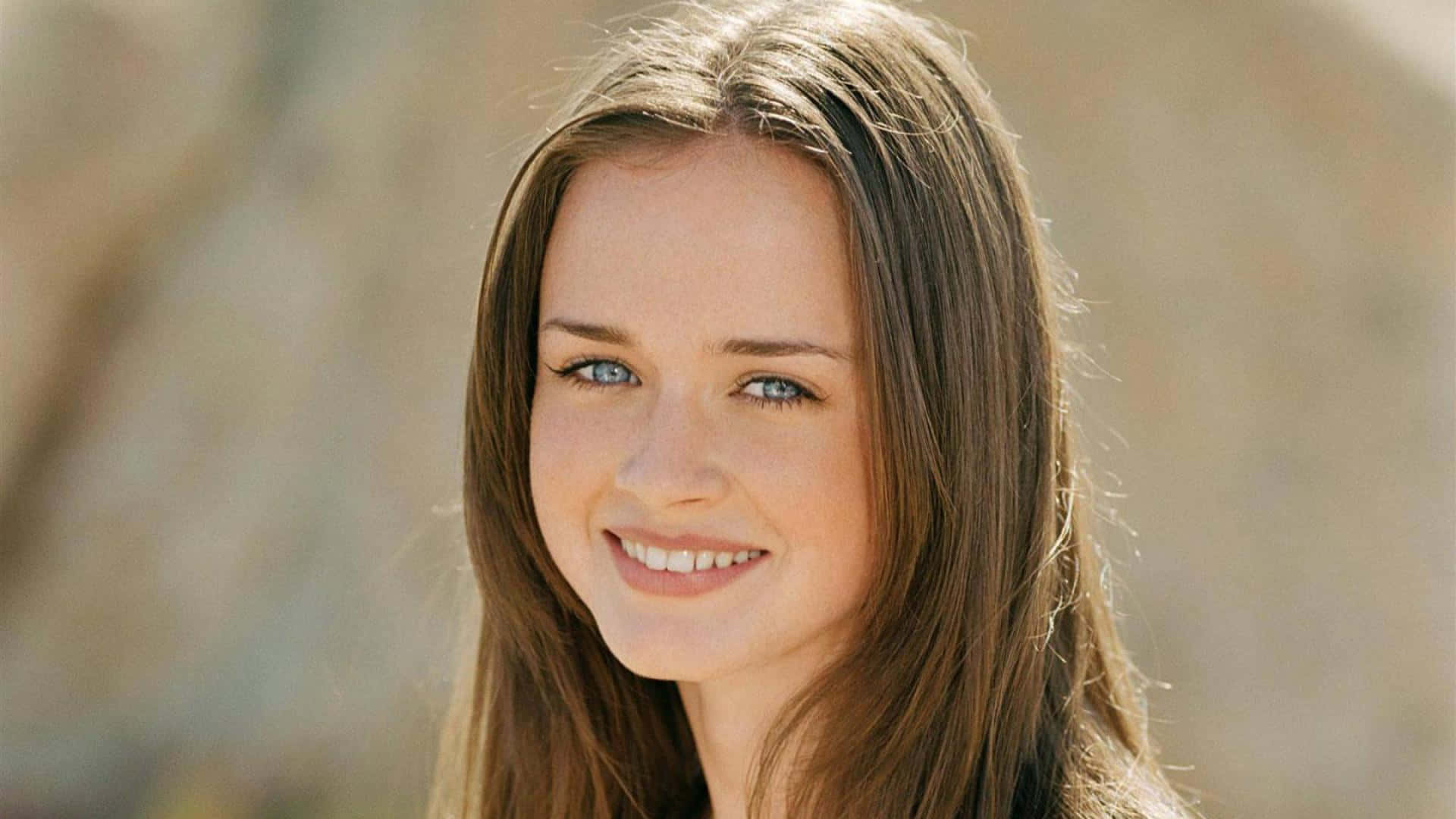 Alexis Bledel 桌布 壁紙 — 一位擁有明亮藍色眼睛和長棕色頭髮的年輕女性，在柔和模糊的自然背景下溫暖地微笑。