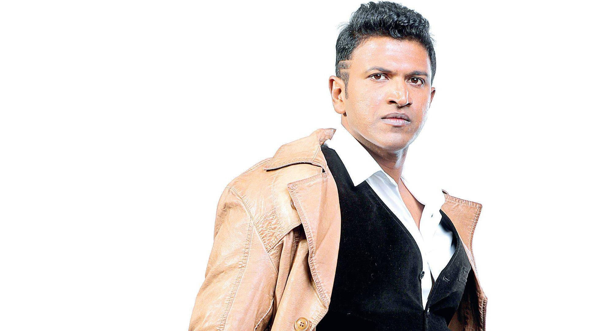 Puneeth Rajkumar 桌布 壁紙 — 一位擁有時尚深色頭髮的男子，在純白背景前穿著棕褐色皮夾克，內搭黑色背心和白色襯衫。