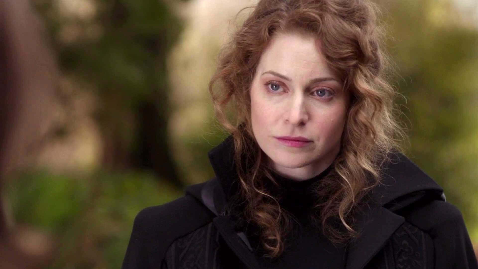 Esmé Bianco 桌布 壁紙 — 一位擁有棕色捲髮和藍色眼睛的女性，在模糊的綠色戶外背景下穿著黑色連帽外套。
