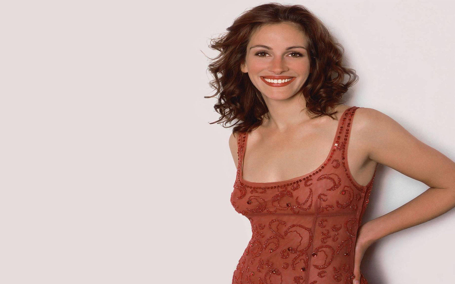 Julia Roberts 桌布 壁紙 — 一位擁有棕色捲髮的女性，身穿紅色珠飾連衣裙，在純白牆壁前溫暖微笑。