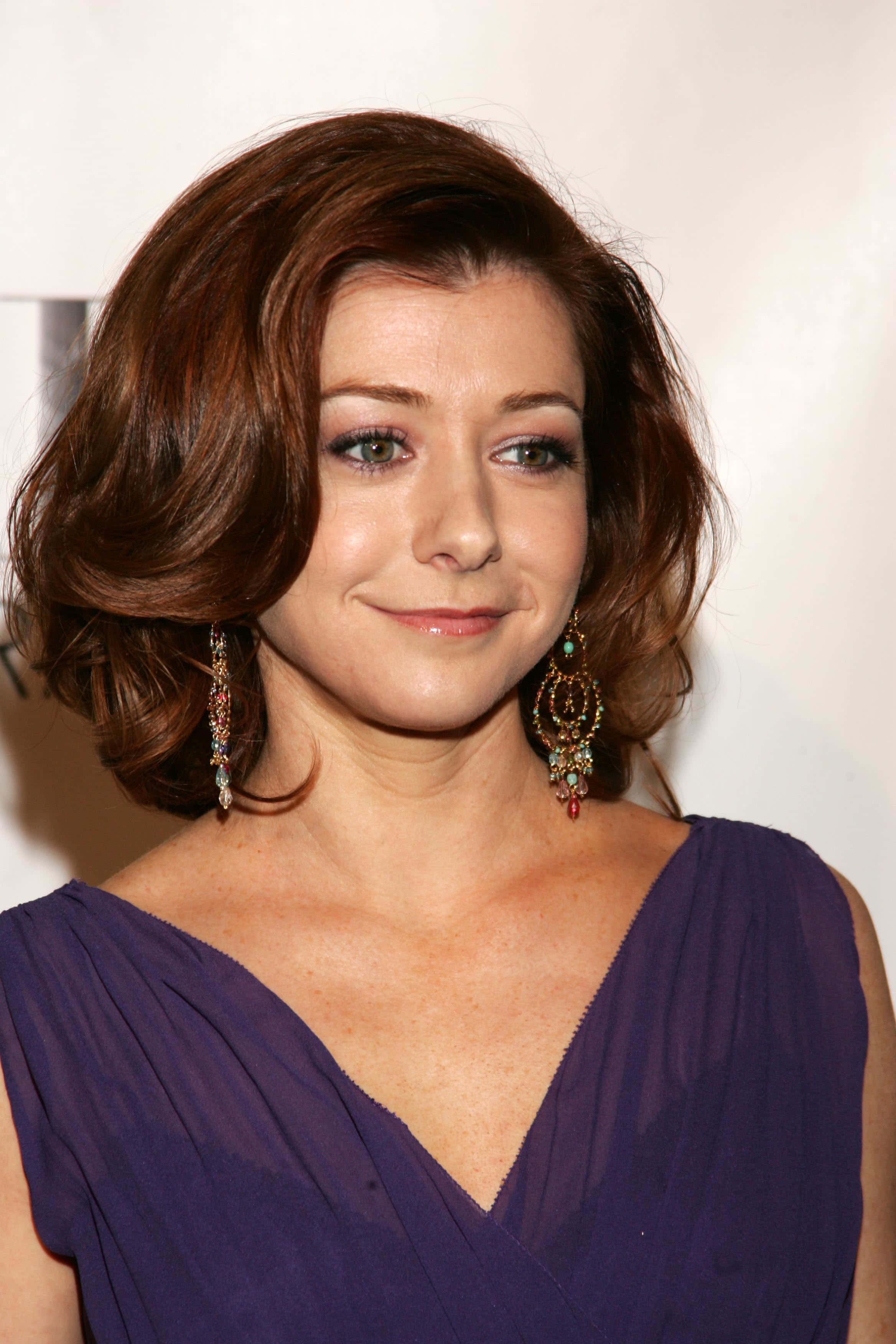 Alyson Hannigan 桌布 壁紙 — 一位擁有棕色波浪長髮和華麗耳環的女性，穿著深紫色垂墜連衣裙，溫柔地微笑。