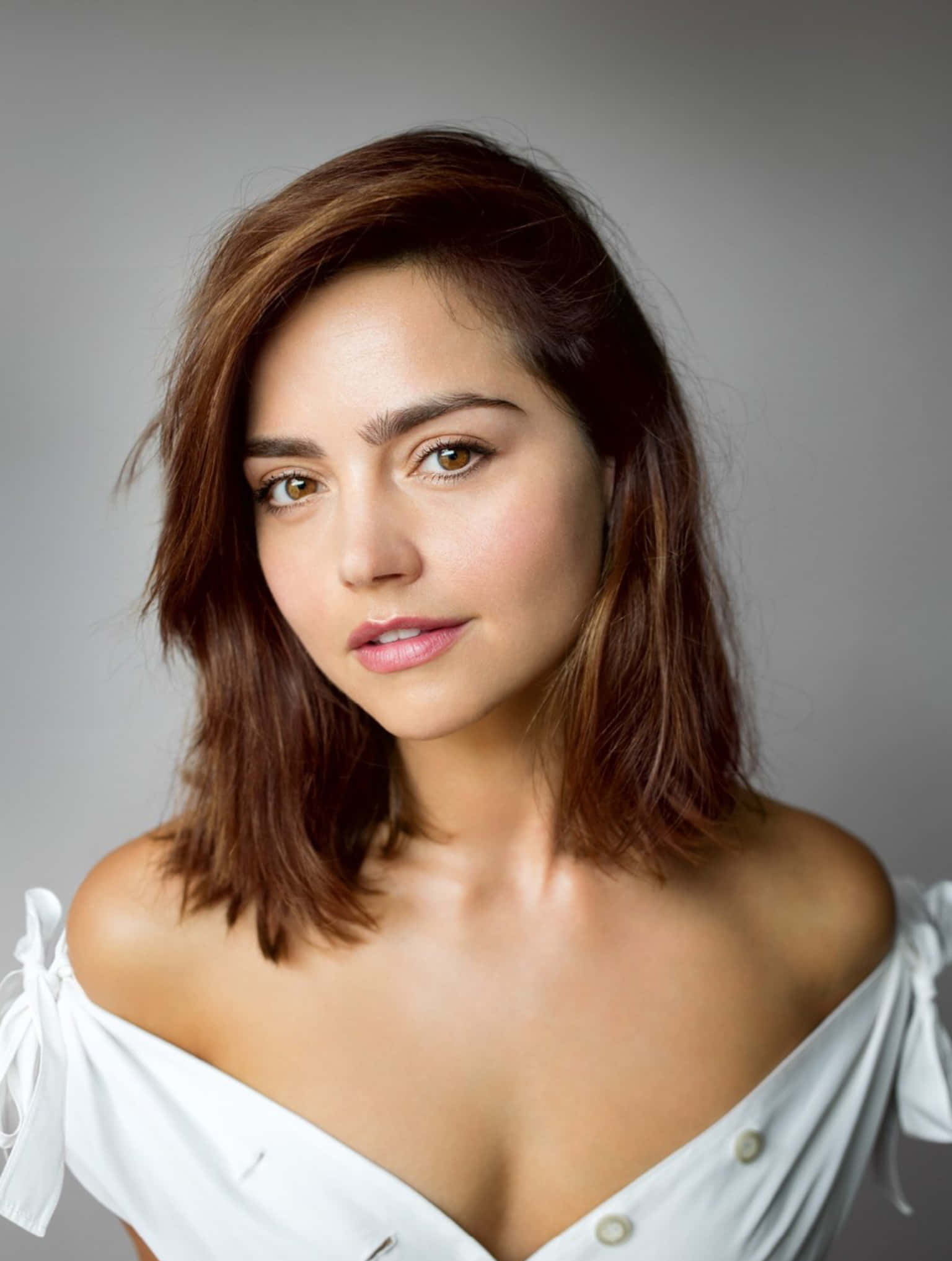 Jenna Coleman 桌布 壁紙 — 一位擁有棕色頭髮和溫暖眼睛的女性，穿著白色露肩上衣，在灰色中性背景前溫柔地凝視。