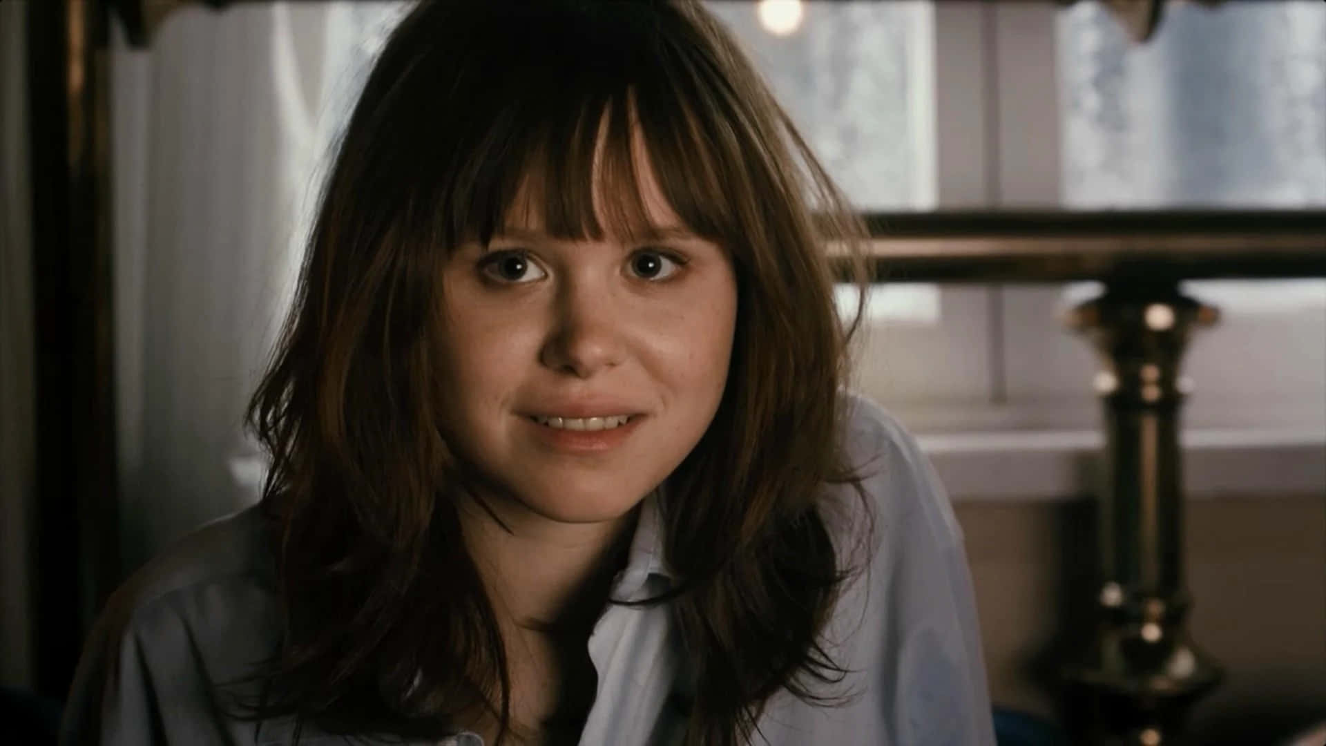 Alison Pill 桌布 壁紙 — 一位擁有棕色頭髮和瀏海的年輕女性，在室內穿著淺藍色襯衫，溫柔地微笑。
