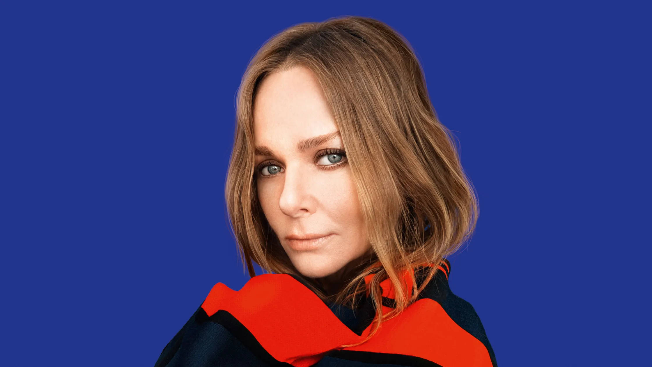 Stella Mccartney 桌布 壁紙 — 一位擁有波浪形棕色頭髮和引人注目的藍色眼睛的女性，在純藍色背景前穿著大膽的紅黑服裝，直視鏡頭。